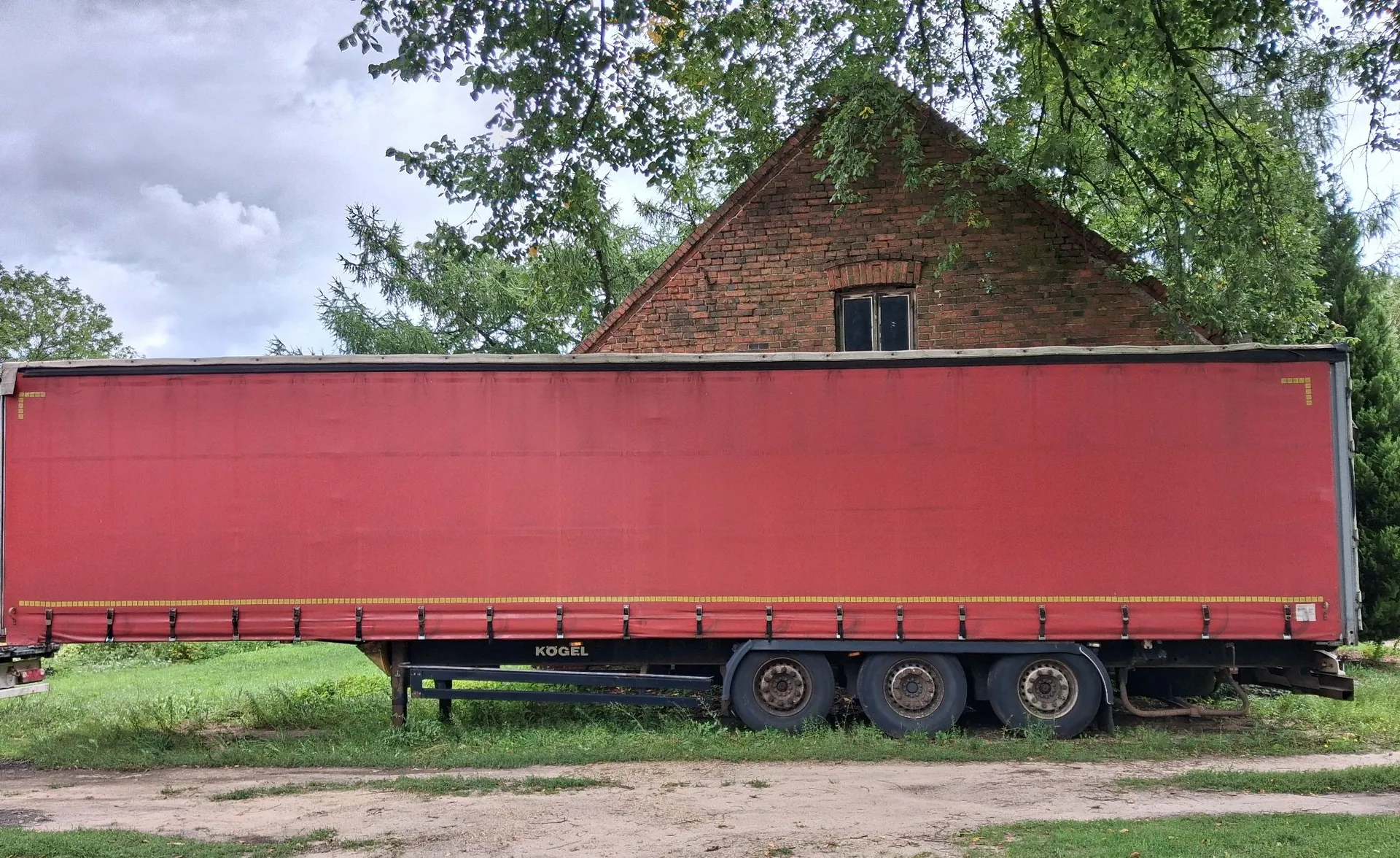 Zdjęcie przedstawiające długą, czerwoną naczepę ciężarówki typu kurtynowego zaparkowaną na trawiastym terenie obok piaszczystej drogi. W tle widoczny jest fragment starego budynku z czerwonej cegły z dwuspadowym dachem oraz zielone drzewa pod zachmurzonym niebem. Na ramie naczepy widnieje napis 'KÖGEL'.