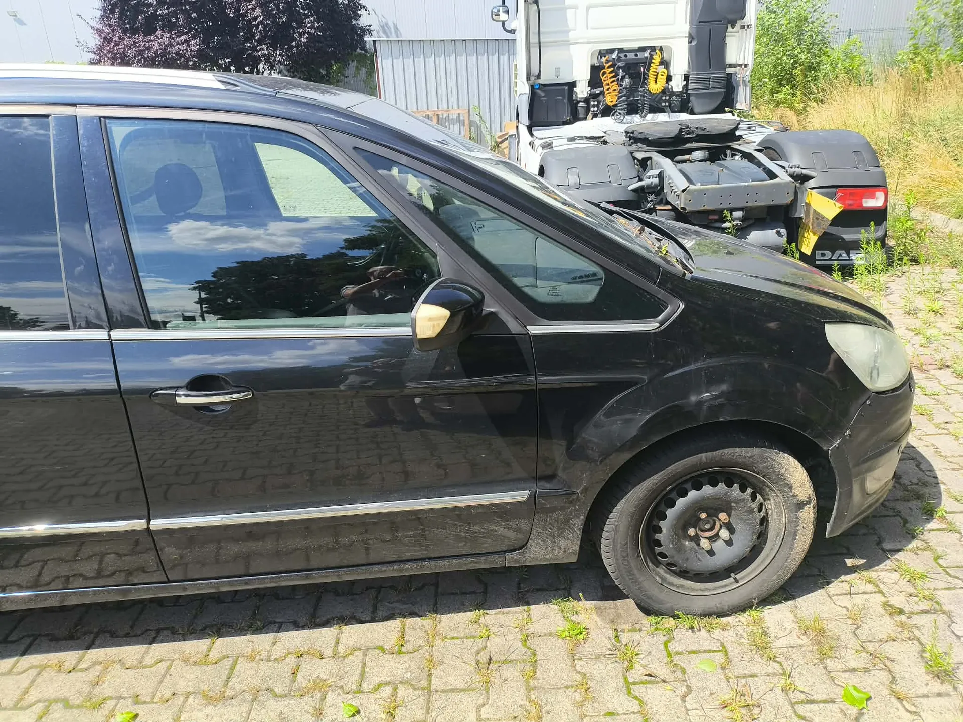 Zbliżenie na prawy bok czarnego samochodu osobowego zaparkowanego na kostce brukowej. W tle widoczny jest biały samochód ciężarowy z platformą lawetową oraz zielone drzewa i krzewy.