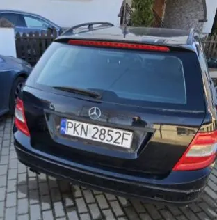 Czarny samochód Mercedes-Benz widziany od tyłu, z tablicą rejestracyjną PKN 2852F. Samochód zaparkowany na brukowanej powierzchni. W tle częściowo widoczny inny pojazd, dom z ogrodzeniem i zieleń. Widoczne są czerwone światła tylnego oraz logo marki na bagażniku.