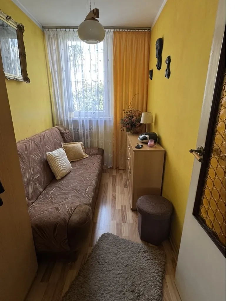 Wnętrze pomieszczenia z żółtymi ścianami. Brązowa sofa z wzorem i dwoma poduszkami (jedna biała, jedna beżowa) po lewej stronie. Okno z białą koronką i pomarańczową zasłoną w tle. Drewniana szafka z lampką, bukietem kwiatów, czarnym urządzeniem i różowym przedmiotem na górze. Okrągły brązowy puf i szary dywan na podłodze z drewnianą parkietową. Na ścianie wiszą czarne dekoracyjne elementy oraz lustro w złotej ramie po lewej. Drzwi z brązową klamką i dekoracyjnym panelem po prawej stronie. Sufit z białą wiszącą lampą.