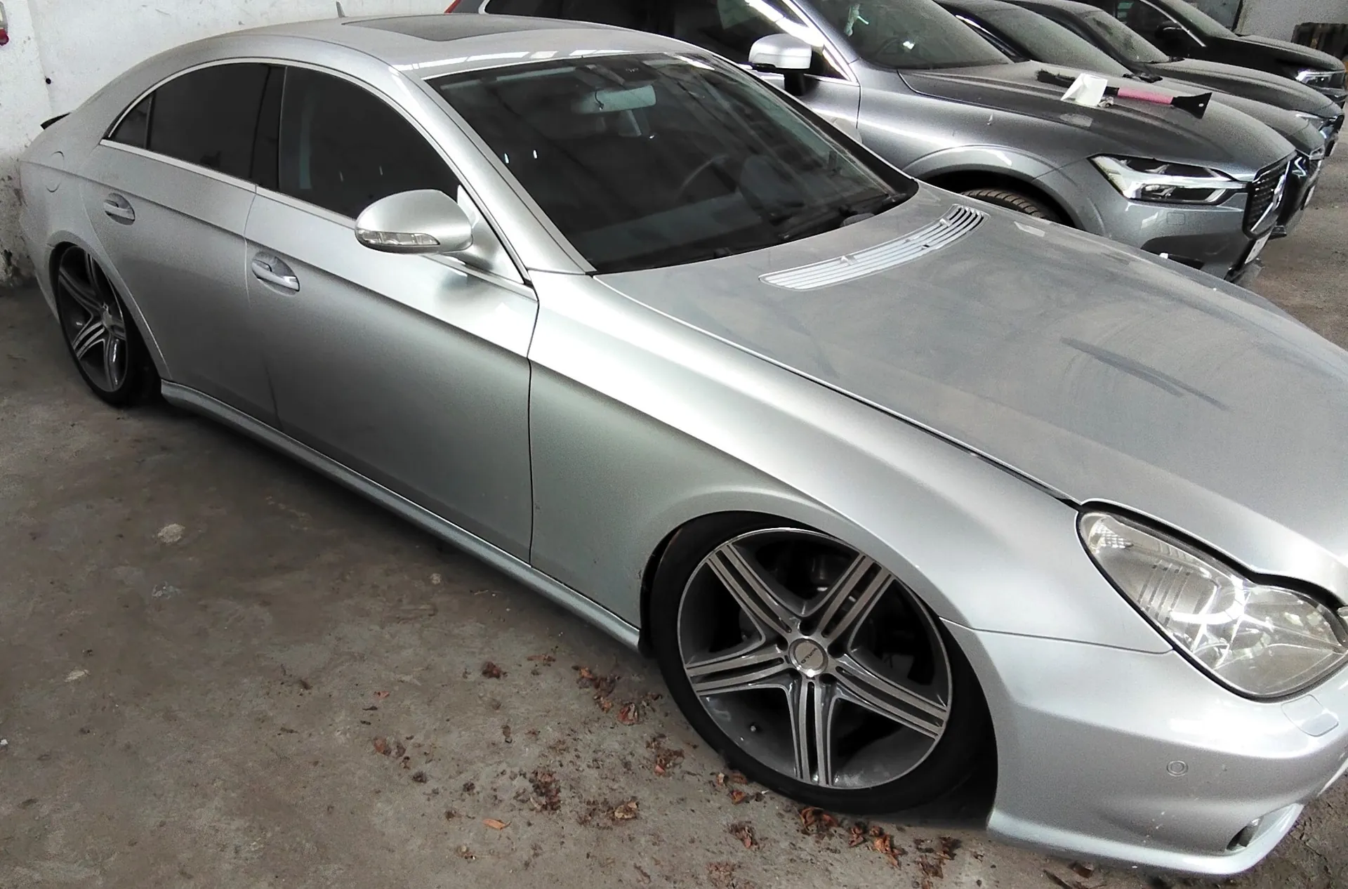 Srebrny samochód osobowy typu coupe zaparkowany w garażu obok innych srebrnych pojazdów. Auto jest widoczne z perspektywy przedniego lewego boku, posiada duże, pięcioramienne felgi aluminiowe.