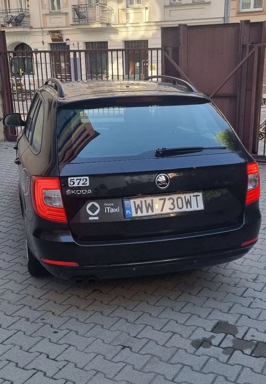 Tył czarnego samochodu marki Skoda zaparkowanego na kostce brukowej. Na klapie bagażnika widnieje logo marki, tablica rejestracyjna o numerze WW 730WT oraz naklejki: '572 ŠKODA' oraz logo 'Grupa iTaxi'. W tle widać brązową bramę oraz fragment jasnej kamienicy.