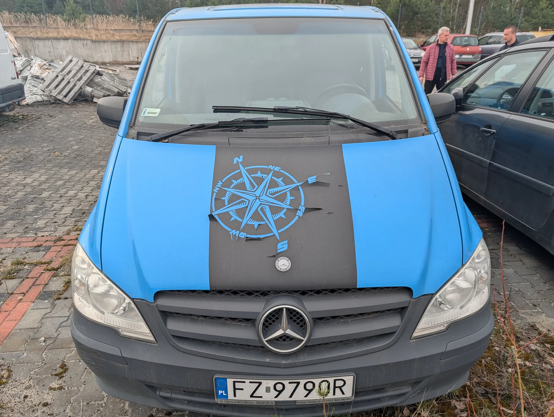 Niebieski samochód dostawczy marki Mercedes-Benz z czarną pionową listwą na masce, na której znajduje się niebieski kompas z oznaczeniami kierunków (N, NE, E, SE, S, SW, W, NW). Tablica rejestracyjna z napisem PL FZ 9790R. W tle widoczne są inne pojazdy, osoba w różowej kurtce, stosy palet i betonowy mur. Podłoga z bruku.