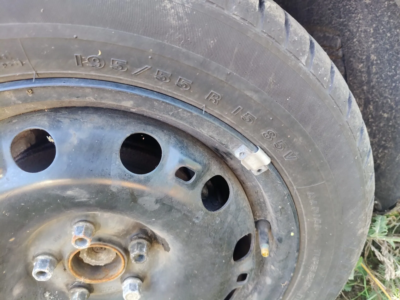 Zbliżenie na czarną stalową felgę samochodową z założoną oponą. Na boku opony widnieje wypukły napis określający rozmiar: 195/55 R 15 85V. Felga posiada kilka okrągłych otworów wentylacyjnych oraz widoczne śruby mocujące.
