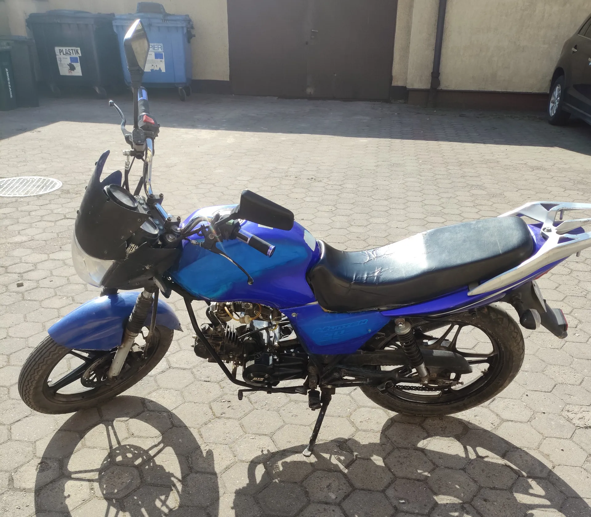 Niebieski motocykl zaparkowany na chodniku z sześciokątnymi płytami. Motocykl ma czarne siedzenie, widoczny silnik i czarny kierownicę z lusterkami. W tle czarne i niebieskie pojemniki na śmieci z napisami "PLASTIK" i "PIER", brązowe drzwi budynku oraz fragment ciemnego samochodu po prawej stronie. Cień motocykla widoczny na chodniku.