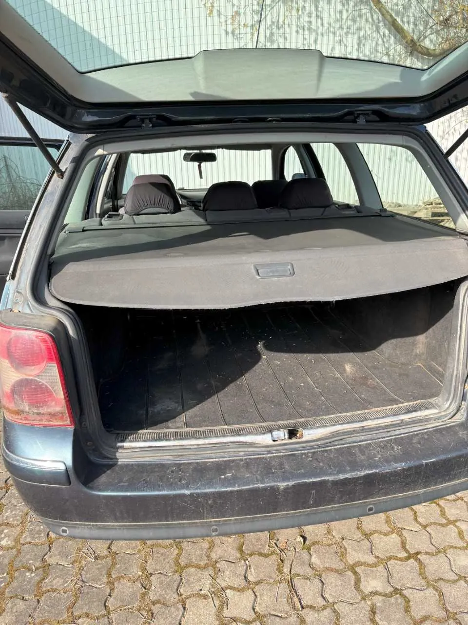 Otwarty bagażnik ciemnoszarego samochodu typu kombi. Wnętrze bagażnika pokryte jest czarną wykładziną, a na środku widoczna jest szara osłona. W tle widać czarne siedzenia, lusterko wsteczne oraz metalowy płot. Samochód stoi na brukowanej powierzchni, a w tle znajdują się drzewa i budynek z metalową ścianą. Na osłonie bagażnika znajduje się niewielki, nieczytelny napis.