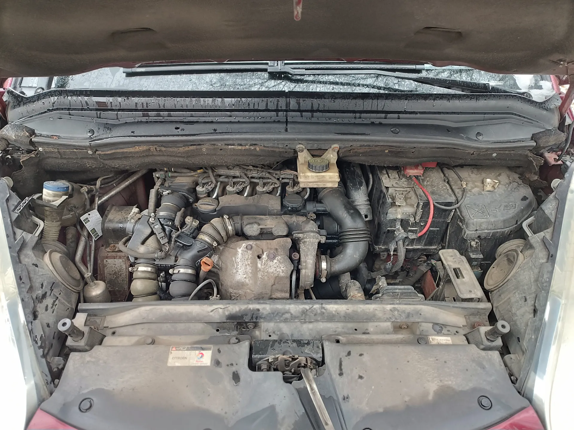 Otwarty kapota samochodu Citroën z widocznym, zanieczyszczonym silnikiem, przewodami, akumulatorem z czerwonym kablem i naklejką „CITROËN”. Na przedniej szybie śnieg, a w tle zimowy krajobraz. Elementy silnika pokryte brudem i kurzem.