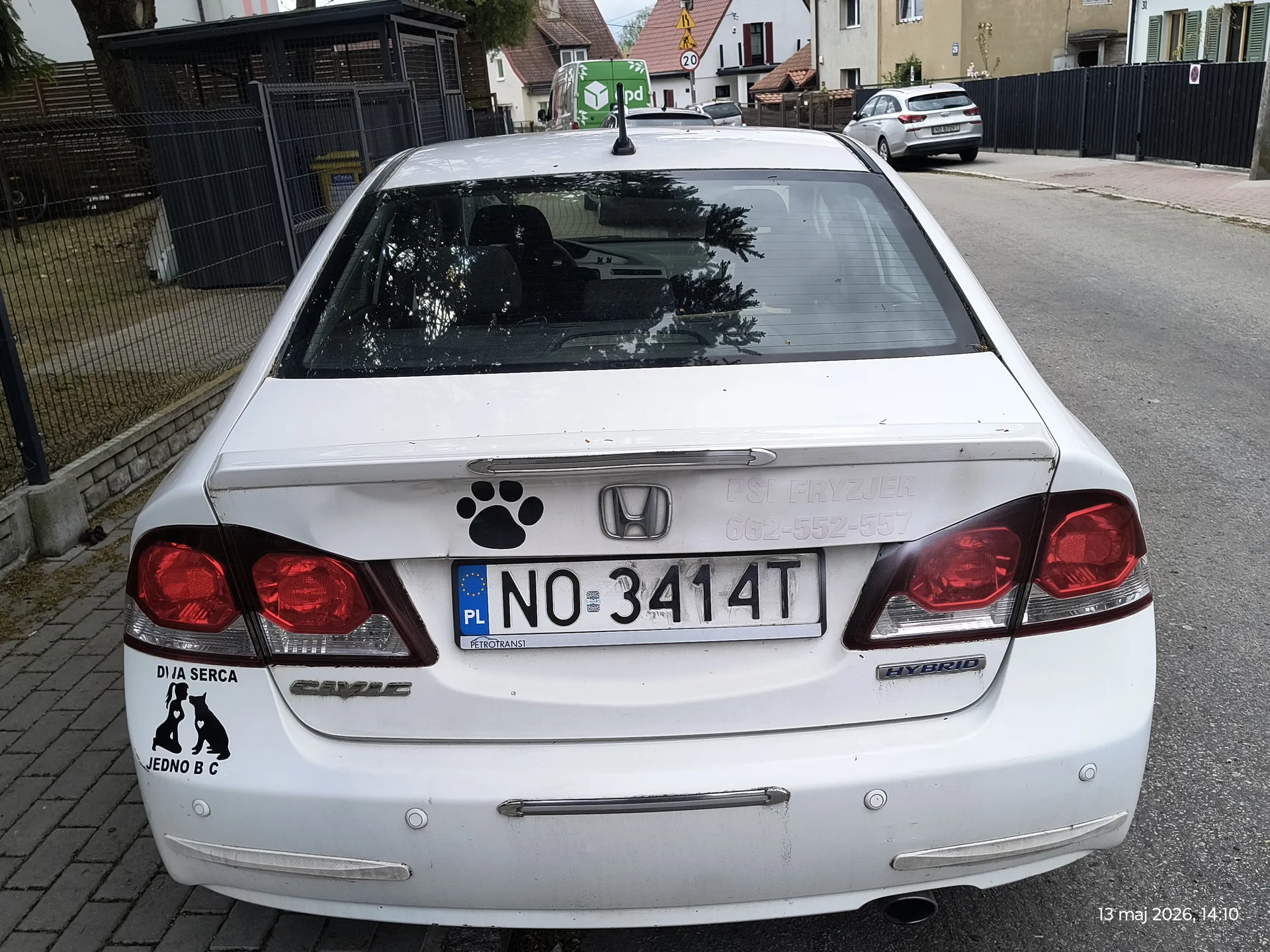Tył białego samochodu Honda Civic Hybrid zaparkowanego na ulicy. Widoczna tablica rejestracyjna z numerem NO 3414T oraz naklejki: czarna łapa, napis 'PSI FRYZJER 662-552-557', a po lewej stronie napis 'DIA SERCA' nad sylwetkami dwóch psów i 'JEDNO B C'. W tle domy, ogrodzenie, zielony furgonetka z logo 'dpd' oraz znak drogowy o ograniczeniu prędkości 20. W prawym dolnym rogu data i godzina: 13 maj 2026, 14:10.