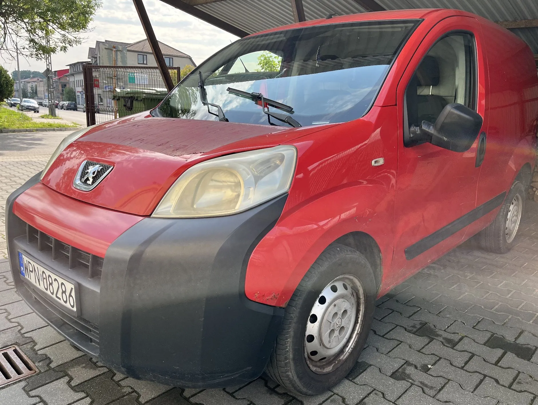 Czerwony samochód dostawczy marki Peugeot zaparkowany pod wiatą na kostce brukowej. Widoczny jest przód i prawy bok pojazdu. Samochód ma czarny zderzak oraz tablicę rejestracyjną o numerze WPN 08286. W tle widać budynki mieszkalne i drzewa.