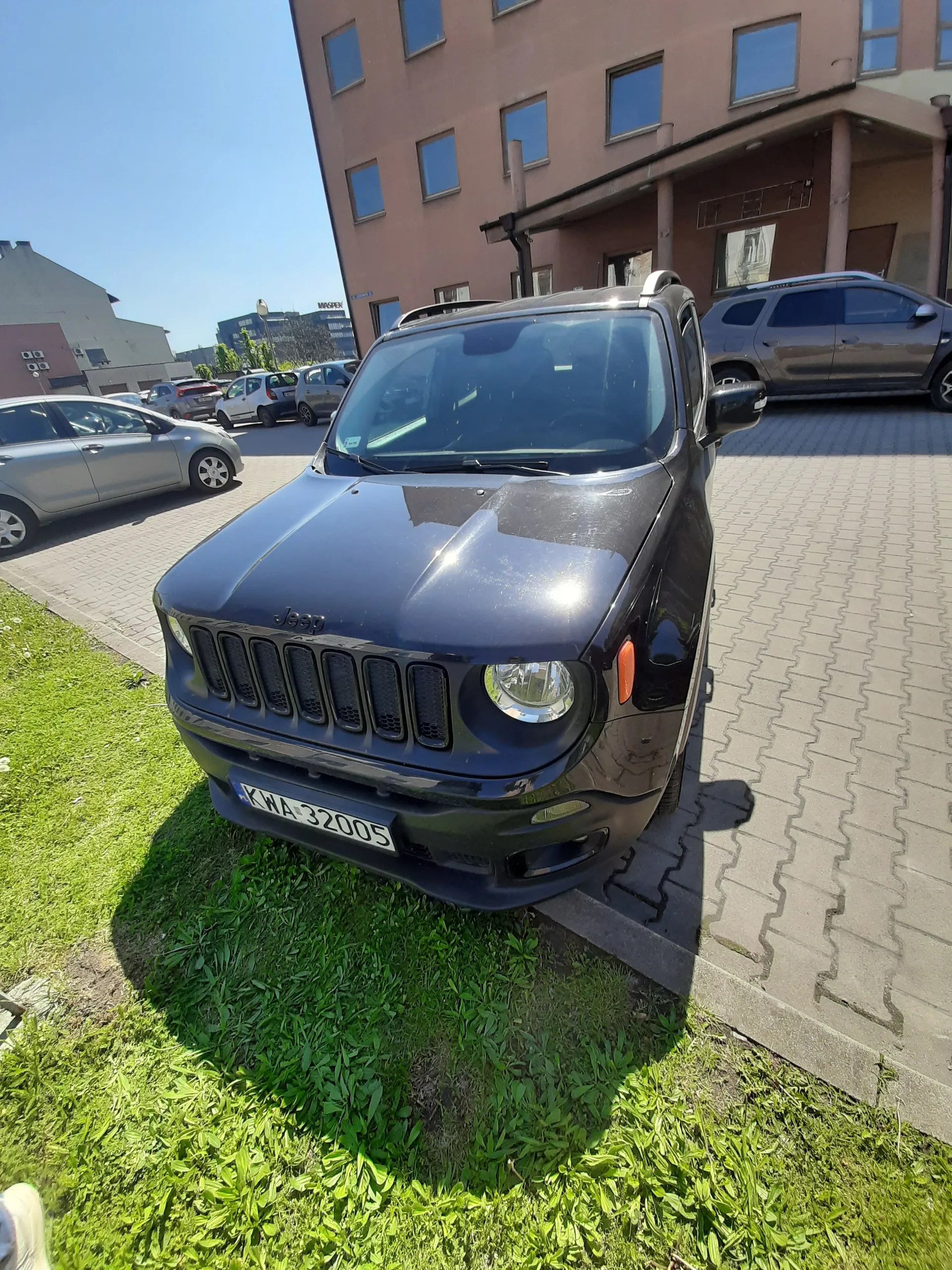 Ciemnoniebieski samochód Jeep z tablicą rejestracyjną KWA 32005 zaparkowany na brukowanej powierzchni obok trawnika. W tle budynki mieszkalne z oknami, inne zaparkowane samochody oraz jasne niebo.