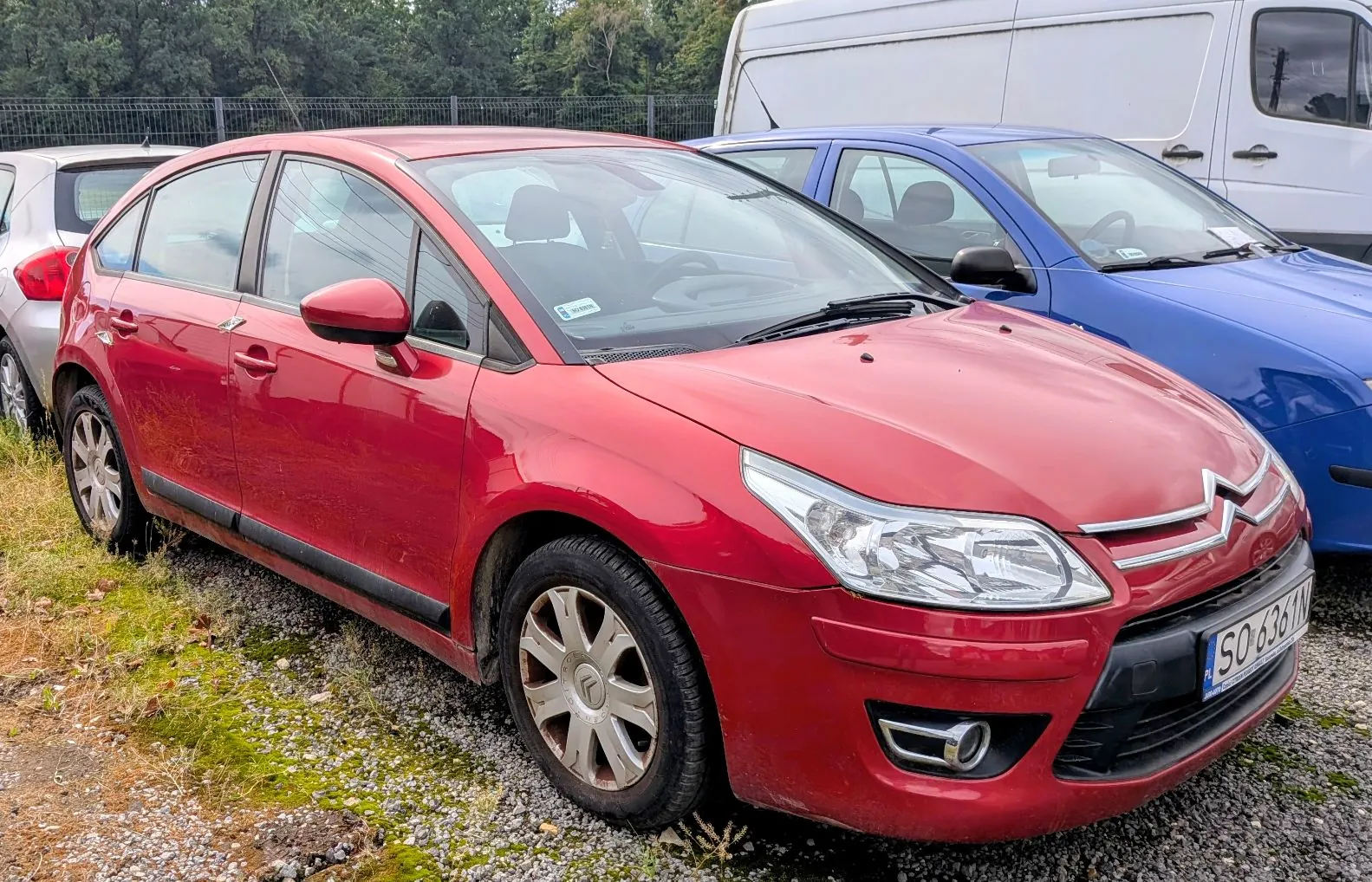 Czerwony samochód osobowy marki Citroën zaparkowany na żwirowym podłożu. Obok niego stoi niebieski samochód, a w tle biały furgon. Samochód ma widoczną tablicę rejestracyjną o numerze SO 6361N.