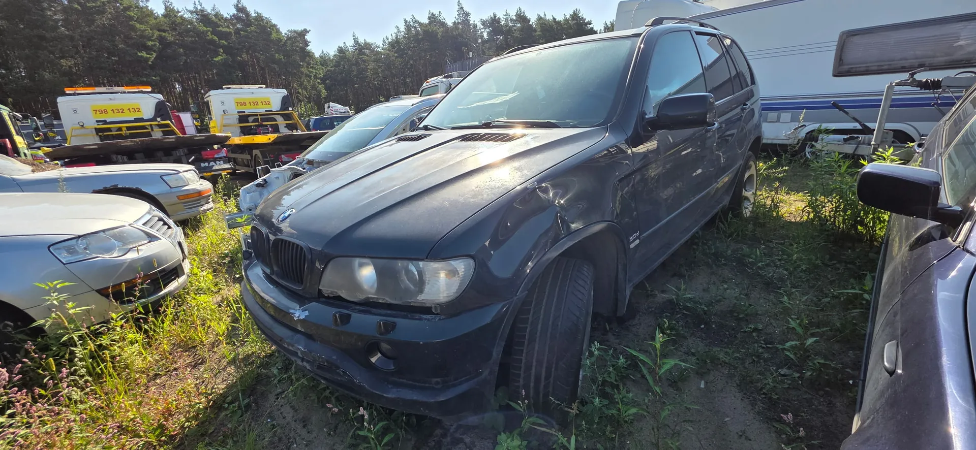 Czarny samochód marki BMW X5 z widocznymi uszkodzeniami karoserii z przodu i z boku, zaparkowany na trawiastym placu. W tle widoczne są inne uszkodzone pojazdy oraz dwa żółto-białe samochody lawety z napisem '798 132 132'. W oddali znajduje się las i biała przyczepa kempingowa.