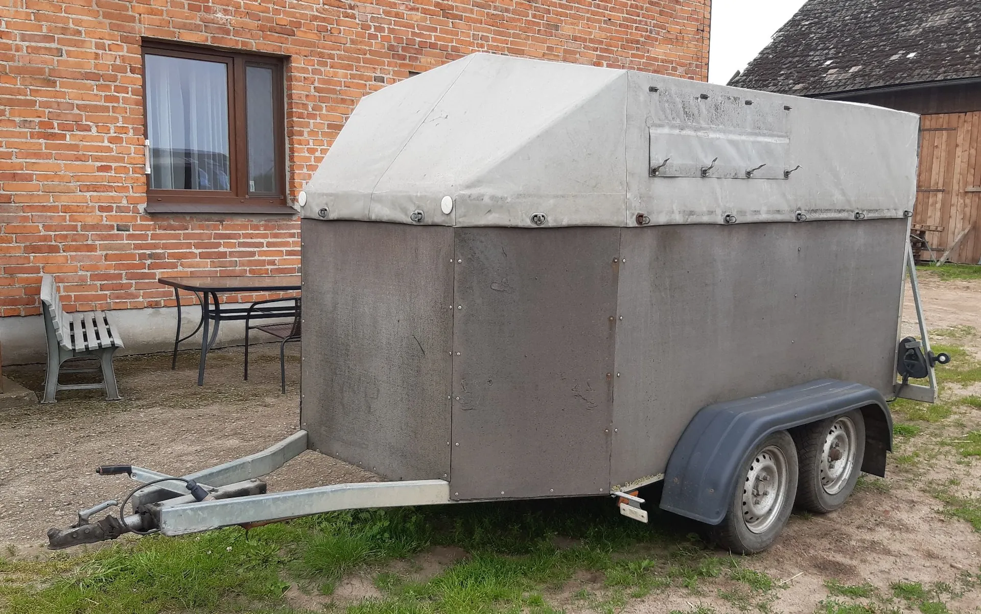 Szara przyczepa towarowa z szarym, płóciennym zadaszeniem, zaparkowana na piaszczystym podłożu przed ceglanym budynkiem. Przyczepa posiada dwie osie z kołami. W tle widoczny jest brązowy stół z krzesłami oraz okno w ceglanej ścianie.