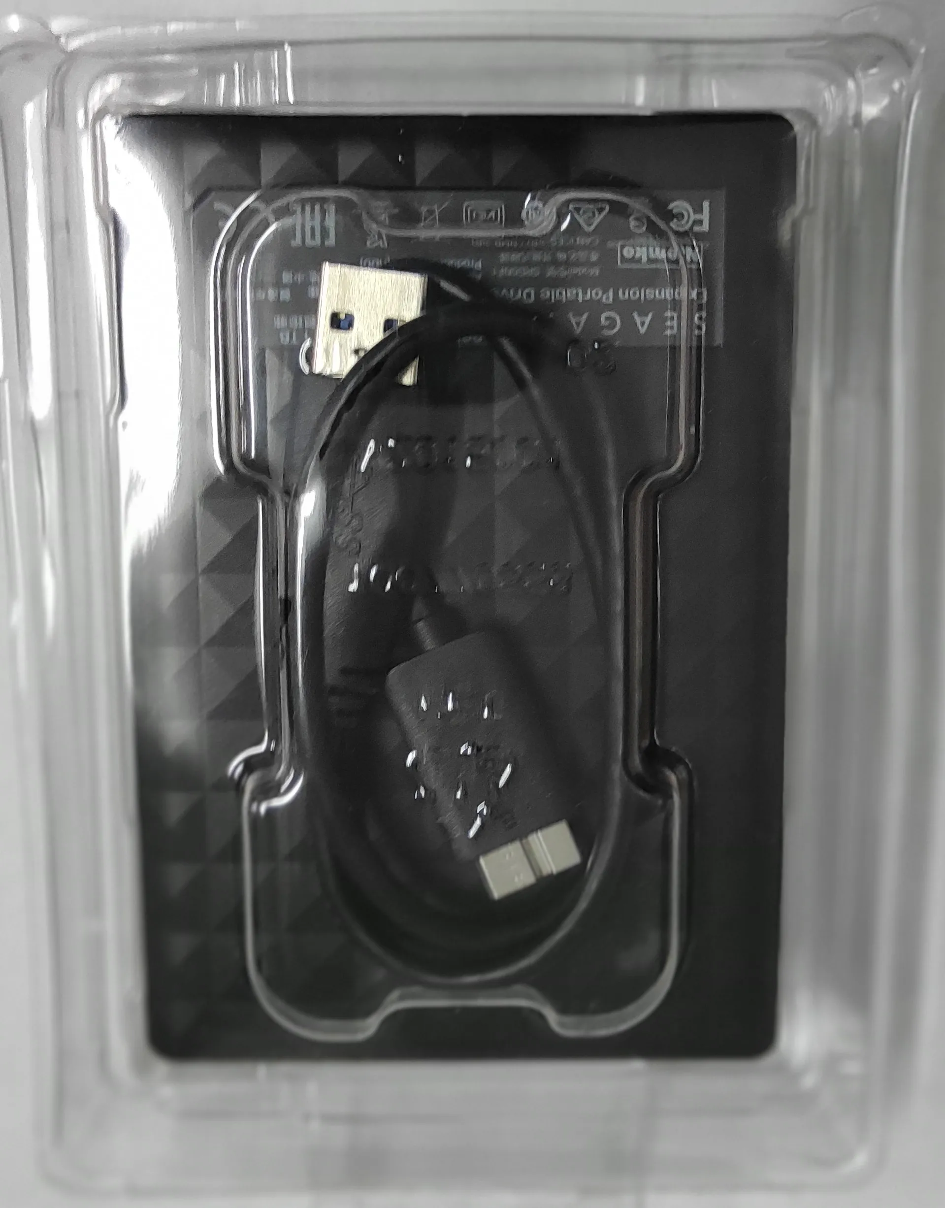 Czarny dysk zewnętrzny marki Seagate Expansion Portable Drive w przezroczystym plastikowym opakowaniu. Do dysku dołączony jest czarny kabel USB, który jest zwinięty i ułożony na wierzchu urządzenia.