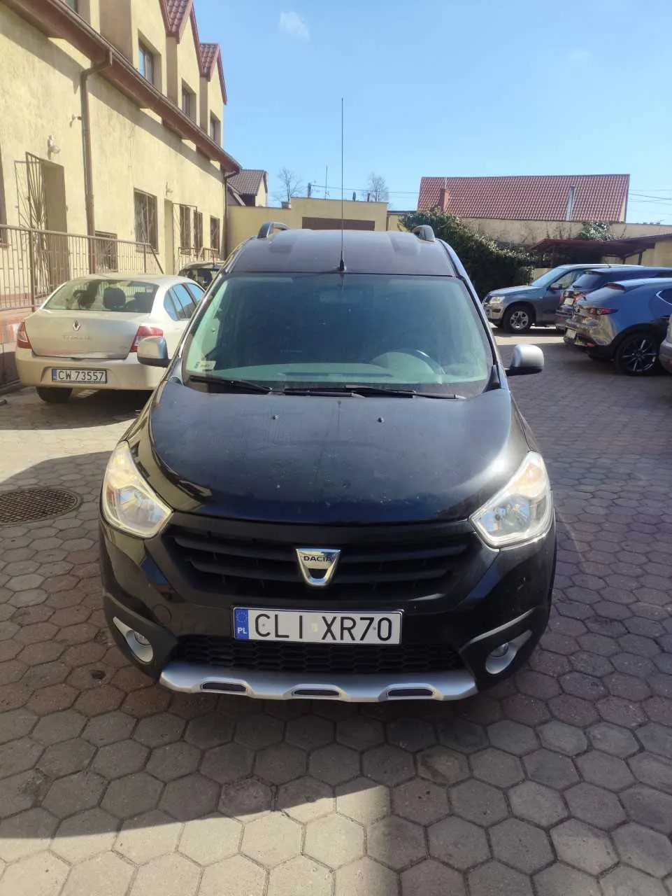 Czarny samochód marki Dacia z tablicą rejestracyjną CL I XR70 na pierwszym planie. W tle widoczne są inne zaparkowane pojazdy, w tym srebrny samochód z tablicą CW 73557, budynki z czerwonymi dachami i jasne niebo. Podłoga wyłożona jest sześciokątnymi płytami brukowymi. Logo Dacia widoczne na przednim zderzaku.