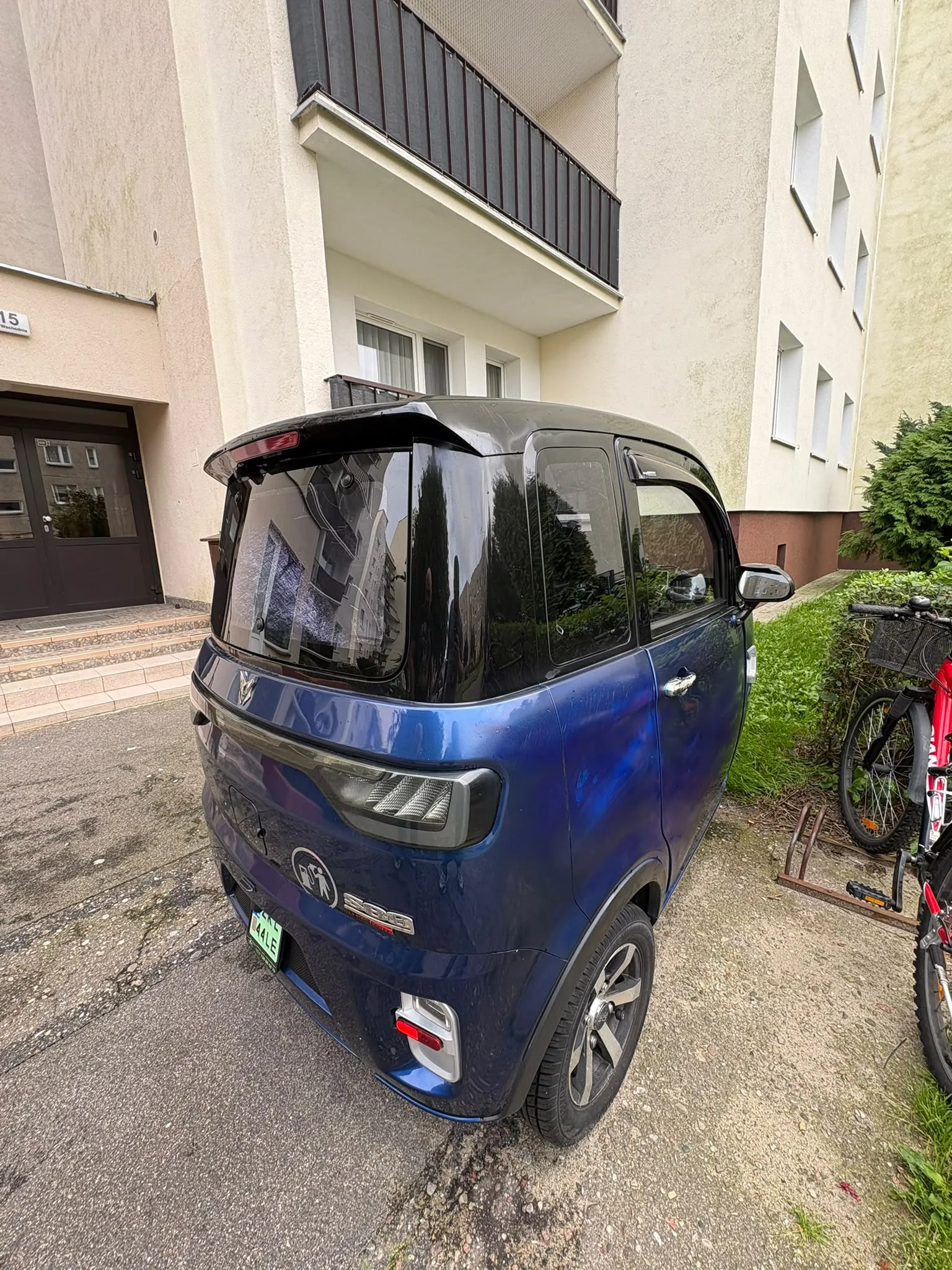 Niebieski, kompaktowy samochód elektryczny zaparkowany na podjeździe przed jasnym, wielopiętrowym budynkiem mieszkalnym. Auto ma ciemny dach, spoiler nad tylną szybą i felgi aluminiowe. Po prawej stronie widoczny jest fragment czerwonego roweru. W tle znajduje się wejście do budynku z ciemnymi drzwiami oraz zielone krzewy.