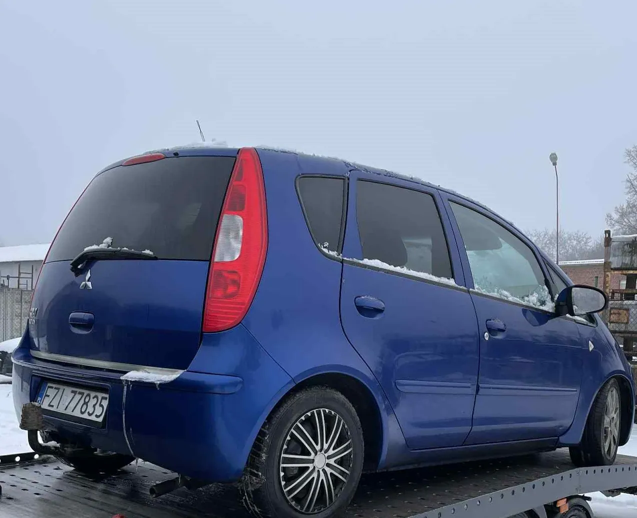 Niebieski samochód osobowy marki Mitsubishi z tablicą rejestracyjną FZI 77835, stojący na platformie holownika. Na dachu i szybach widoczny jest śnieg. Tło przedstawia zimowy krajobraz z budynkami, latarnią uliczną i bezlistnymi drzewami. Samochód ma czarne felgi i czerwone światła tylnego. Niebo jest pochmurne.