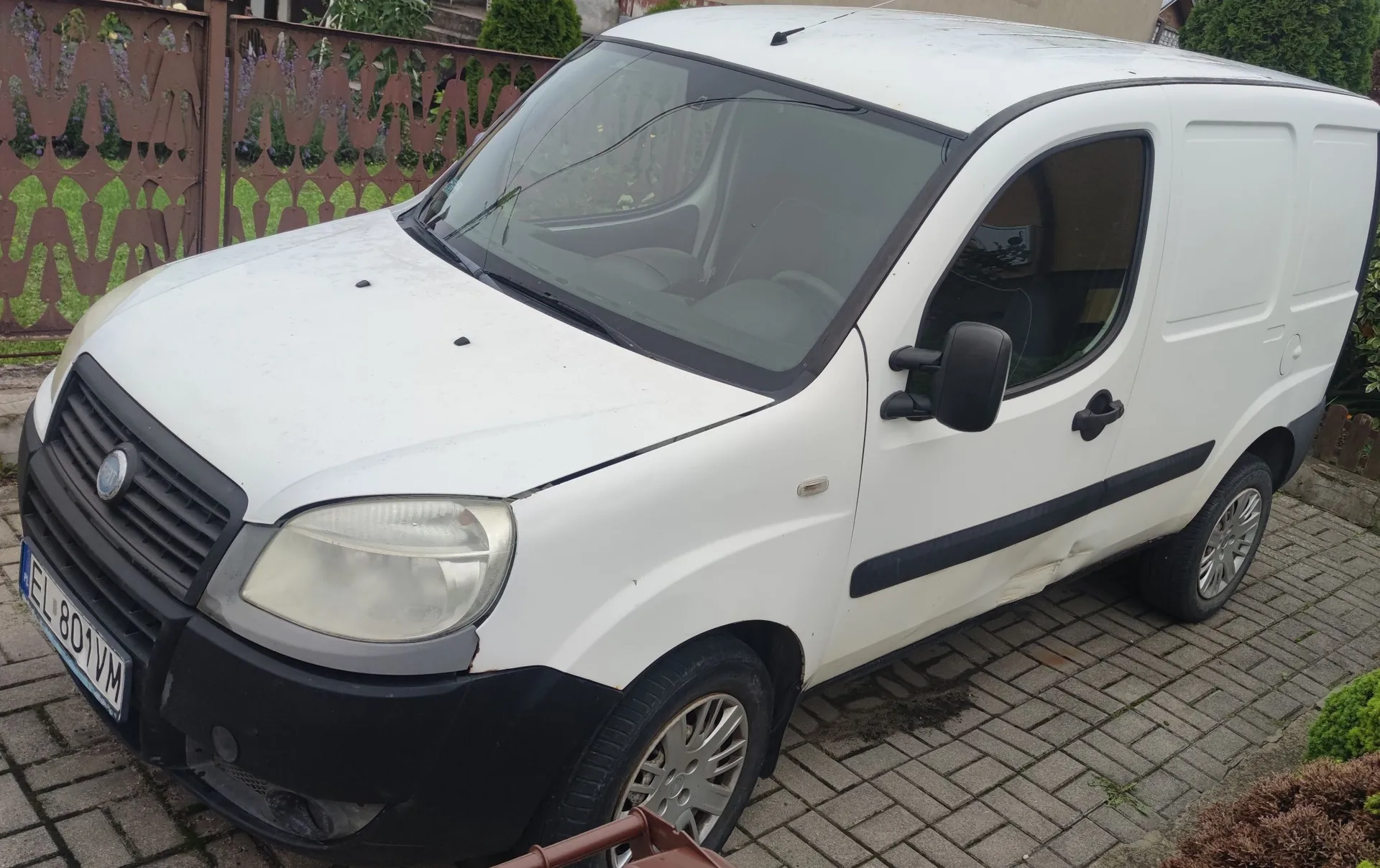 Biały samochód dostawczy typu furgon zaparkowany na kostce brukowej. Pojazd ma czarny zderzak przedni, czarne listwy boczne oraz polską tablicę rejestracyjną o numerze EL 801VM. W tle widoczny jest brązowy metalowy płot z ozdobnymi wycięciami oraz zieleń ogrodowa.