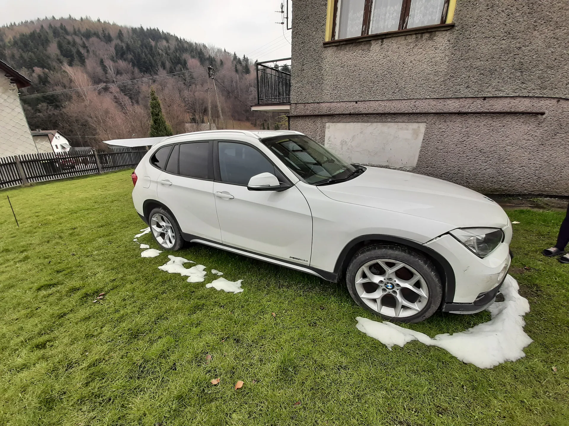 Biały samochód typu SUV zaparkowany na zielonej trawie z resztkami śniegu. W tle widoczne są budynki mieszkalne, ogrodzenie, las oraz wzgórza. Po prawej stronie obrazu częściowo widoczna jest noga osoby w czarnych spodniach i butach. Niebo jest zachmurzone.
