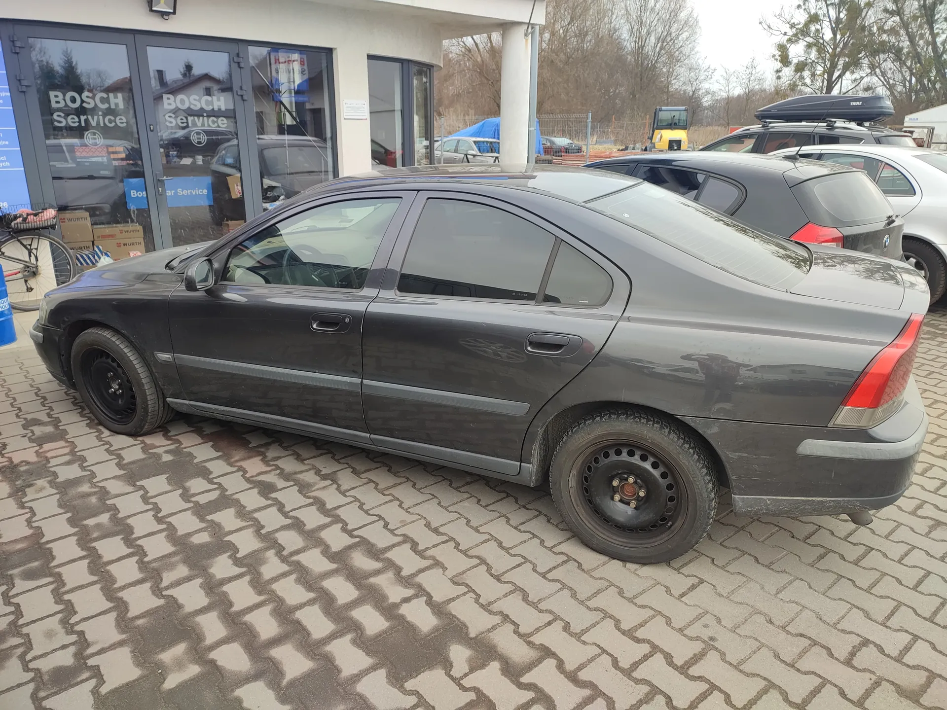 Ciemnoszary samochód osobowy typu sedan zaparkowany na brukowanej powierzchni przed budynkiem z napisem "BOSCH Service" na szklanych drzwiach. W tle widoczne są inne pojazdy, m.in. czarny hatchback i srebrny samochód, a także rower, pudełka z napisem "WURTH" oraz drzewa bez liści. Budynek ma białą elewację z dużymi oknami, a na tle widać niebo i maszynę budowlaną.