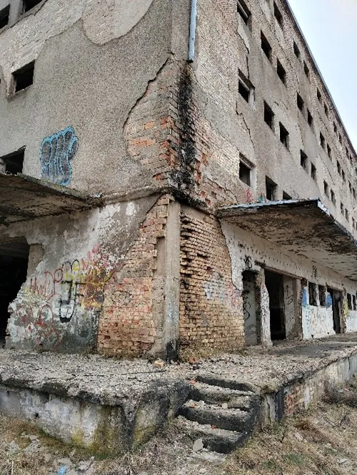 Zdjęcie przedstawia róg zniszczonego, wielopiętrowego budynku z cegły i betonu. Tynk na ścianach odpada płatami, odsłaniając czerwoną cegłę. Na ścianach widoczne są liczne graffiti w kolorach niebieskim, czerwonym i żółtym. Budynek posiada rzędy małych, prostokątnych okien bez szyb. Przed wejściem znajduje się betonowy podest z kilkoma zniszczonymi stopniami schodów. Teren wokół jest nieuporządkowany, z widoczną suchą trawą i gruzem.