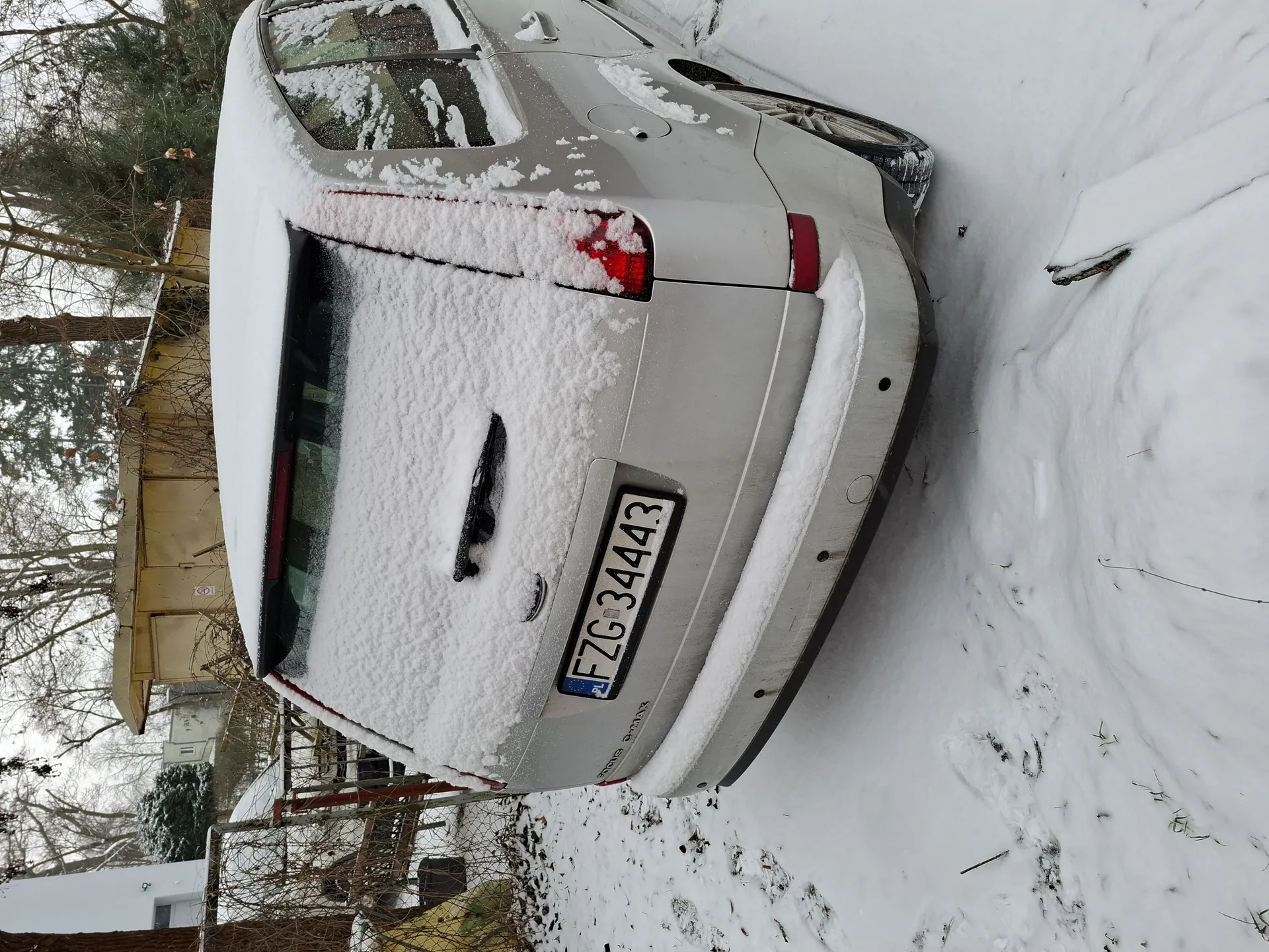 Srebrny samochód pokryty śniegiem, widok od tyłu. Tablica rejestracyjna FZG 34443 z niebieskim pasem z napisem PL po lewej stronie. Śnieg pokrywa tylne szyby, dach i fragmenty nadwozia. W tle żółty domek, drzewa bez liści, ogrodzenie oraz częściowo widoczny inny samochód. Śnieżna powierzchnia ziemi z odciskami stóp i rozproszonymi gałązkami.