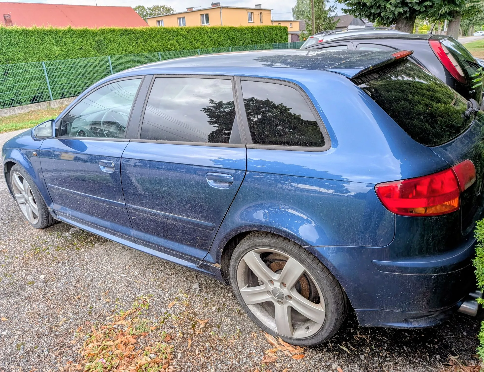 Niebieski samochód typu kombi zaparkowany na żwirowym podjeździe. Widoczny jest profil auta z boku, z naciskiem na tylną część i koła z pięcioramiennymi felgami aluminiowymi. W tle znajduje się zielony żywopłot, niebieski płot oraz żółty budynek mieszkalny pod czerwonym dachem.