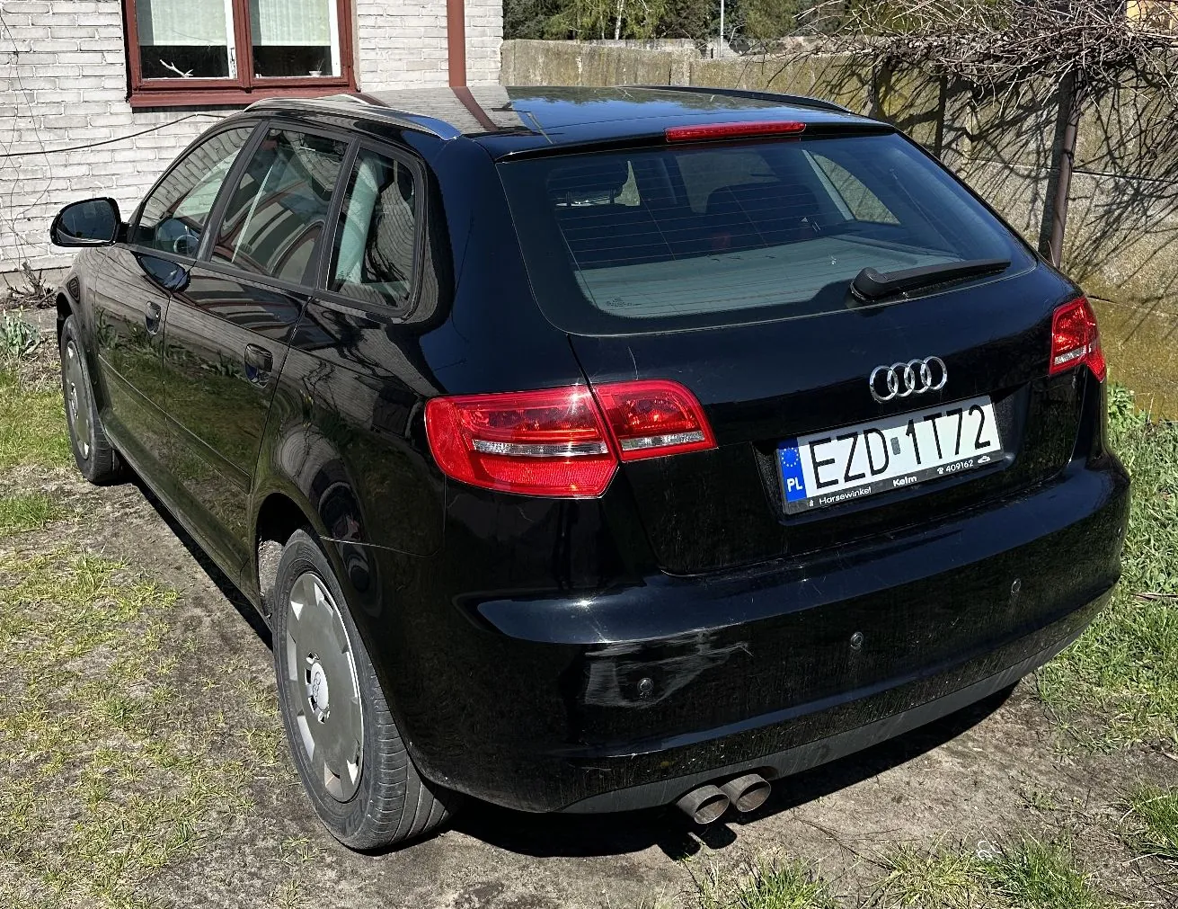 Czarny samochód marki Audi w nadwoziu kombi, zaparkowany na trawiastym podłożu przed jasnym budynkiem. Widoczny jest tył i lewy bok pojazdu. Samochód posiada polską tablicę rejestracyjną o numerze EZD 1T72.