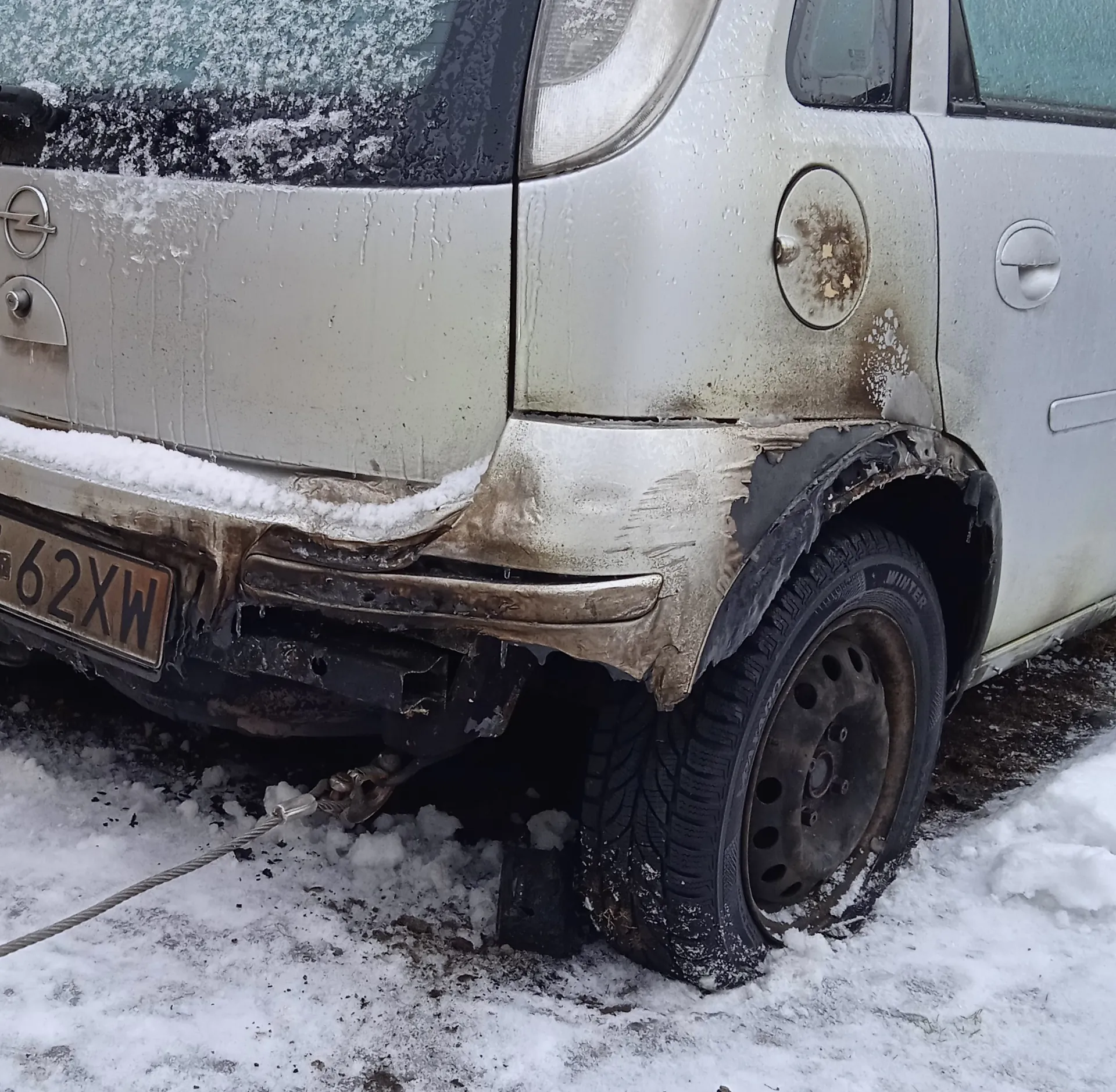 Zdjęcie przedstawia tylną część srebrnego samochodu z widocznymi uszkodzeniami. Błotnik i zderzak są spalone lub stopione, a na powierzchni pojazdu znajdują się plamy rdzy. Na samochodzie widoczny jest śnieg, szczególnie na przedniej szybie i nadkolu. Tablica rejestracyjna z numerem 62XW jest umieszczona po lewej stronie. Do tyłu pojazdu przytwierdzony jest linowy holownik. Opona na tylnej osi to opona zimowa z widocznym napisem WINTER. Podwozie i koło pokryte są śniegiem oraz brudem. Tło stanowi śnieżna powierzchnia z widocznymi śladami.