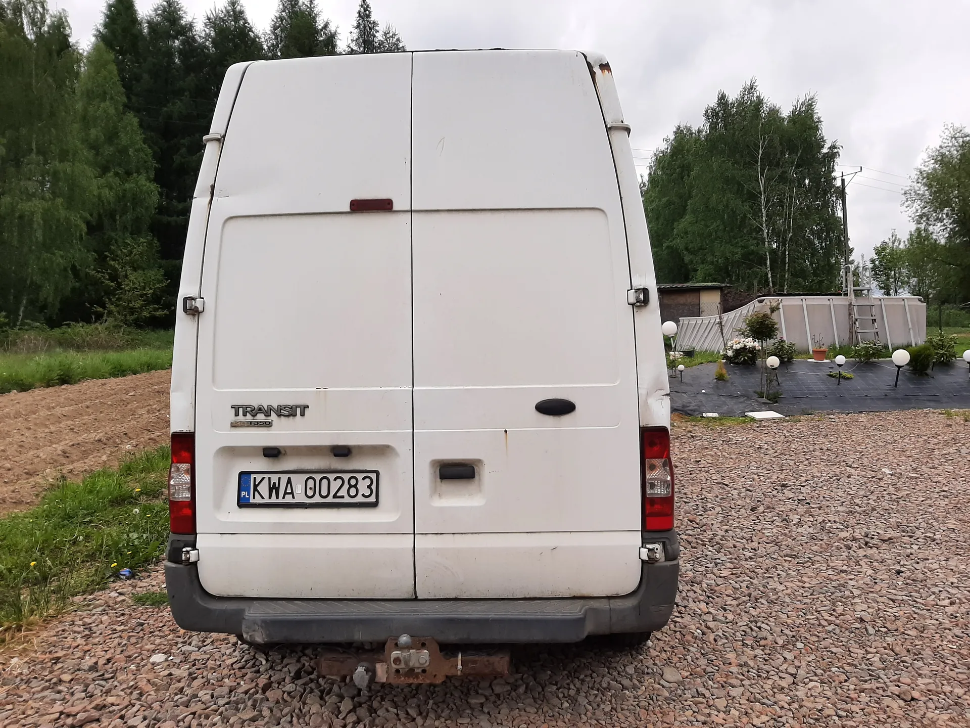 Tył białego furgonu marki Transit z tablicą rejestracyjną KWA-00283 (PL) na żwirowym podłożu. W tle drzewa, zielona trawa, ogrodzenie, struktura przypominająca basen z drabiną oraz kuliste latarnie. Na furgonie widoczne są czerwone światła tylna, napis TRANSIT oraz ślady rdzy w górnym prawym rogu. Niebo jest zachmurzone.