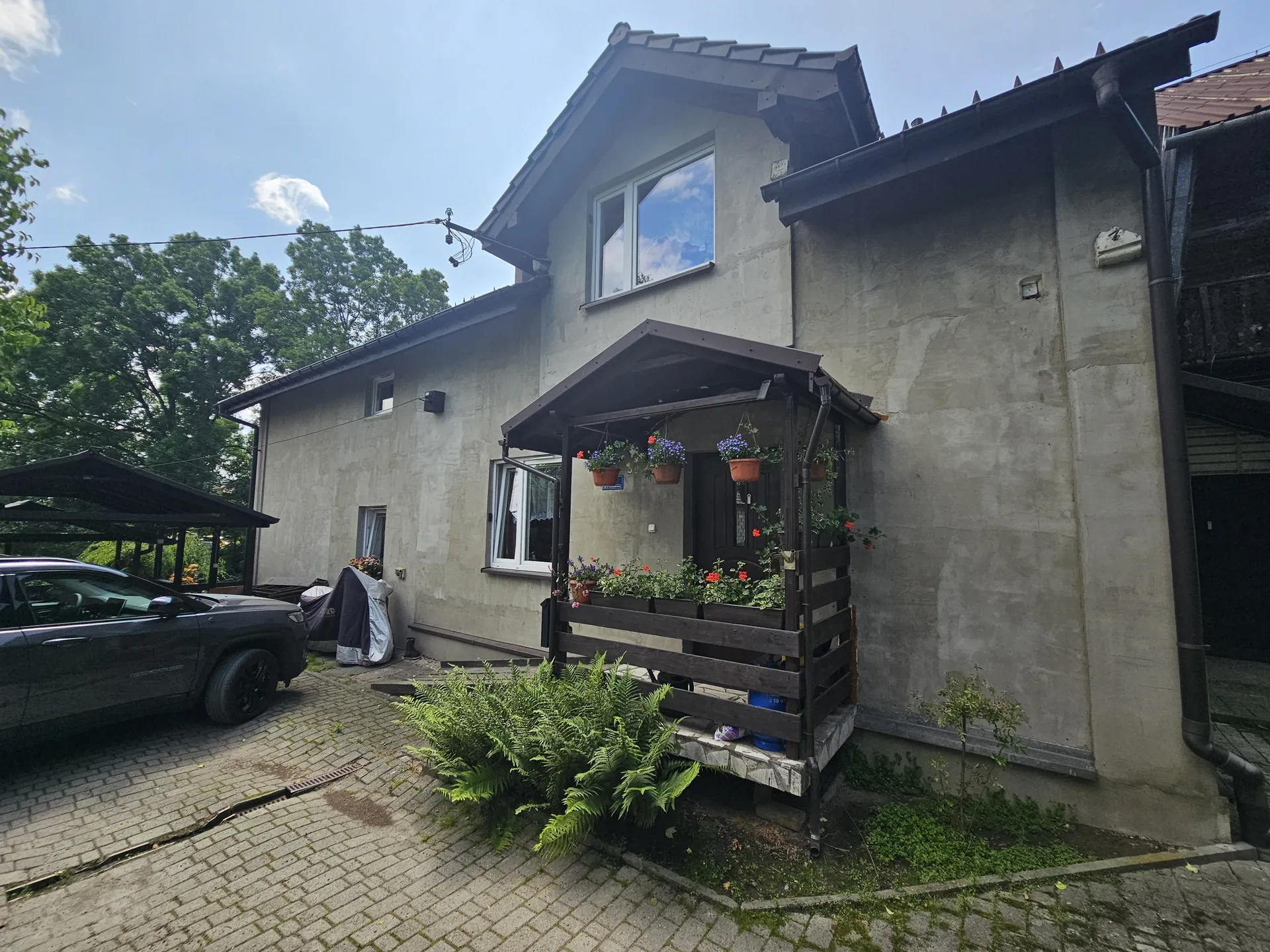 Dwupiętrowy dom o betonowej elewacji z daszkiem nad wejściem, pod którym wiszą doniczki z kwiatami. Przed domem brukowana podjazdowa, na której stoi ciemny samochód osobowy. Po lewej stronie widoczny namiot garażowy oraz zakryte przedmioty. Wokół domu rosną krzewy i drzewa, na niebie kilka chmur. Okna domu są zamontowane w standardowej pozycji, horyzont jest poziomy.