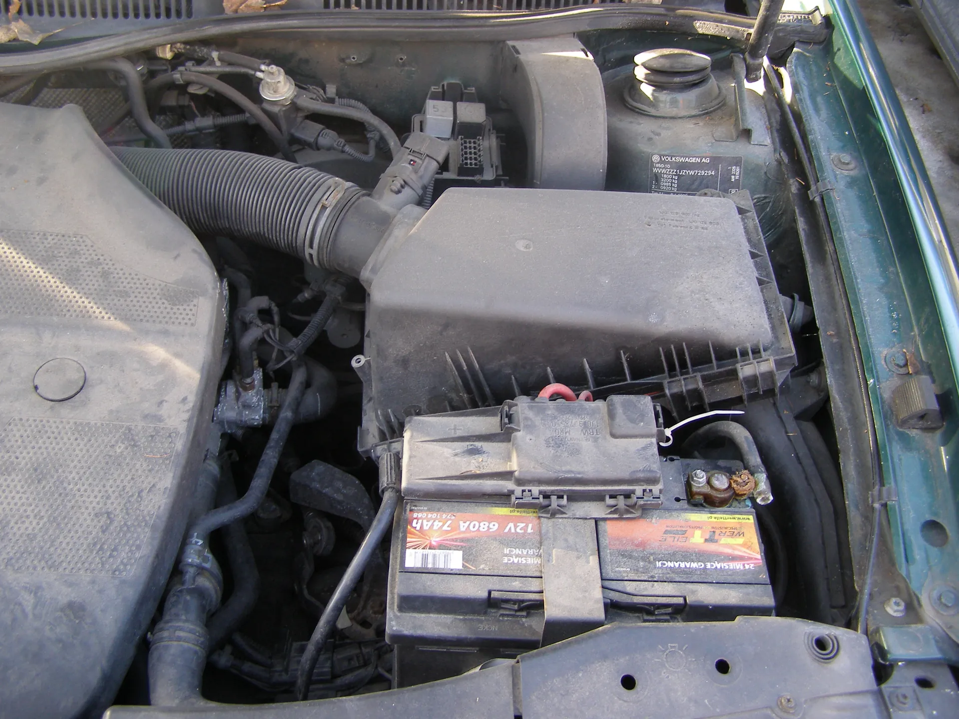 Widok otwartej tylnej części silnika samochodu marki Volkswagen. W centrum znajduje się czarna bateria z widocznymi etykietami: '12V 680A 74Ah', 'WARTA', 'MIEJSCA GWARANCJI' oraz kodem kreskowym. Nad baterią umieszczony jest czarny plastikowy pojemnik z napisem 'VOLKSWAGEN AG' oraz numerem seryjnym. Wokół widoczne są czarne przewody, węże, w tym gruby, spiralny wąż powietrzny. Po lewej stronie znajduje się szary, perforowany element osłony silnika. Po prawej stronie widoczna jest zielona krawędź nadwozia samochodu. Całość pokryta jest kurzem i zabrudzeniami.