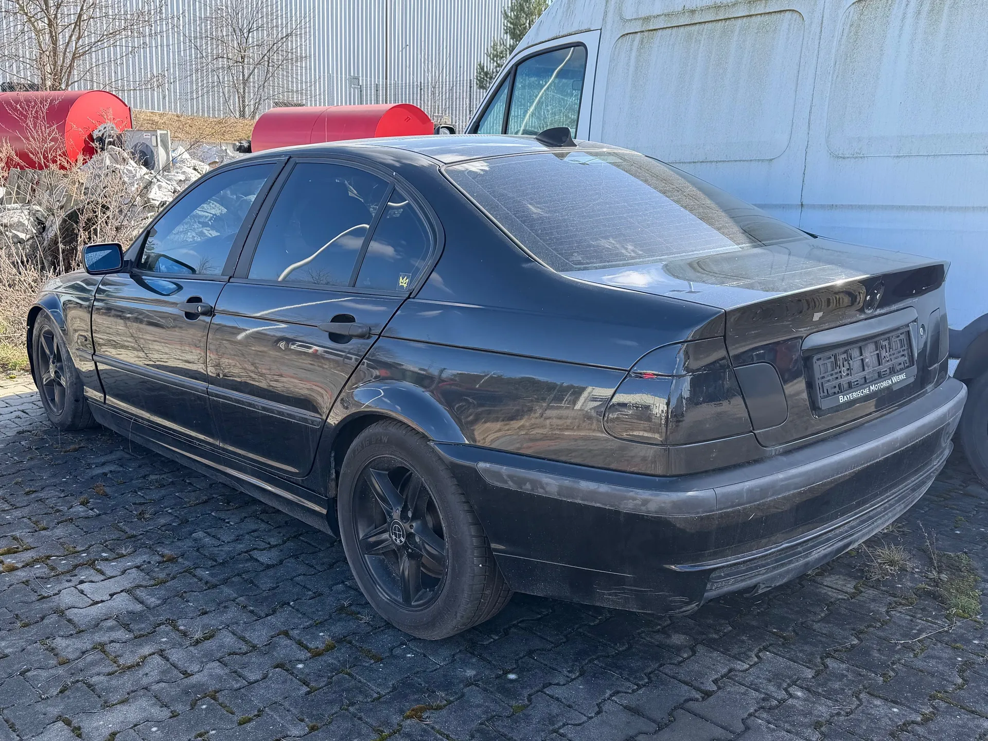 Czarny samochód osobowy typu sedan zaparkowany na brukowanej powierzchni. Widok z tyłu i boku. Po prawej stronie biały furgon. W tle czerwone zbiorniki, metalowe ogrodzenie oraz suche krzewy. Na tablicy rejestracyjnej widoczny napis 'Bayerische Motoren Werke'.