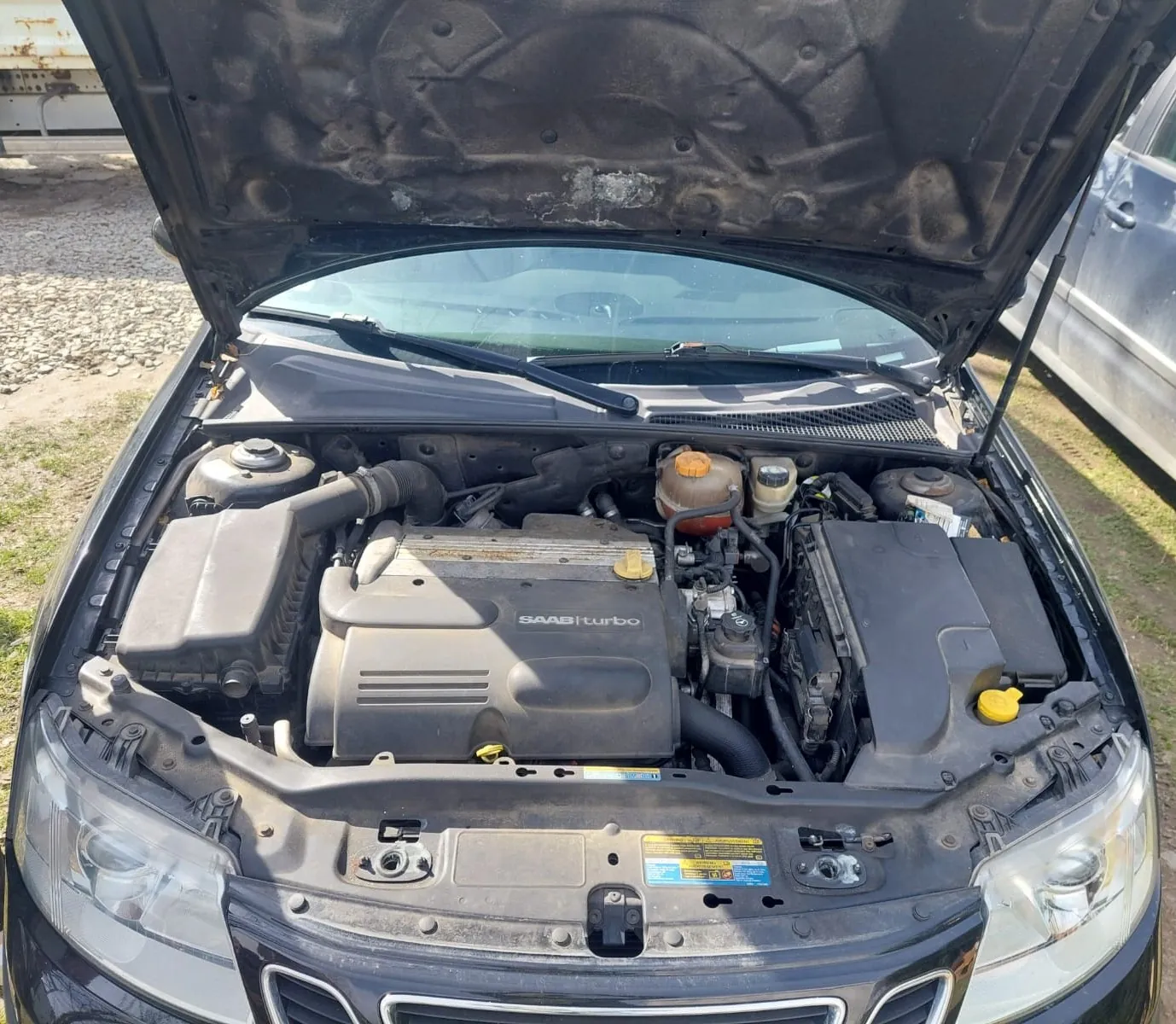 Widok z góry na otwartą maskę czarnego samochodu, ukazującą komorę silnika. Na osłonie silnika widnieje napis 'SAAB turbo'. Widoczne są poszczególne elementy mechaniczne, w tym zbiornik płynu chłodniczego, akumulator oraz przewody. W dolnej części zdjęcia widoczny jest fragment przedniego grilla i reflektorów samochodu.
