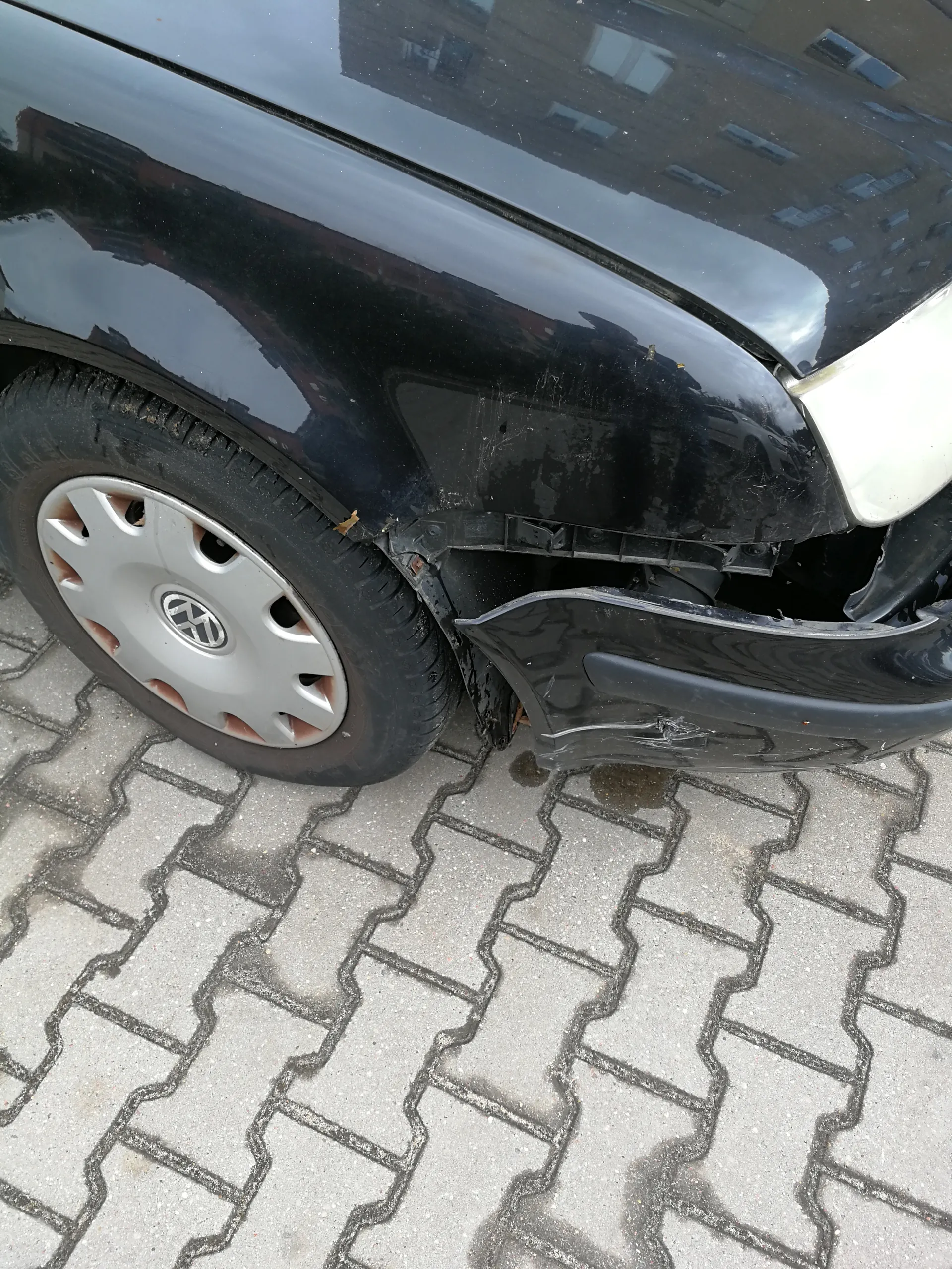 Czarny samochód marki Volkswagen z uszkodzonym przodem po prawej stronie. Widoczne zdeformowane zderzak i błotnik, odsłonięte elementy podwozia. Koło z srebrną felgą z logo VW. Samochód zaparkowany na chodniku z betonowych płyt w kształcie litery S. W odbiciu na karoserii widać budynki z oknami.