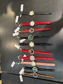 Wiele zegarków na rękę ułożonych w rzędzie na ciemnej powierzchni. Zegarki mają różne kolory pasków: brązowy, różowy, czerwony, czarny. Każdy zegarek posiada białą etykietę. Tarcze zegarków różnią się wzorami i kolorami, w tym białe, zielone i inne.