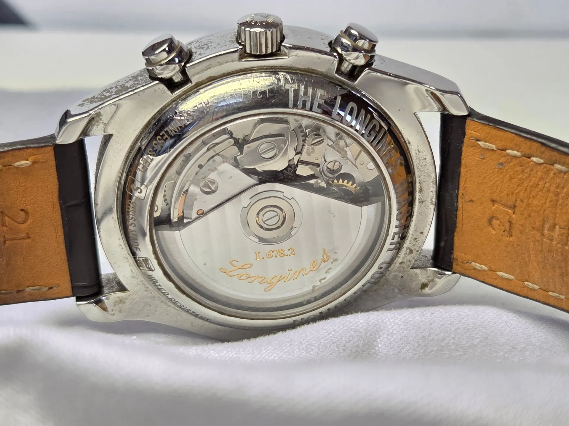 Zbliżenie na tył zegarka marki Longines z przezroczystym deklem, ukazującym mechanizm automatyczny. Na szklanym dnie widnieją napisy: 'L678.2' oraz duże logo 'Longines'. Metalowa koperta posiada trzy pokrętła u góry i jest przymocowana do brązowego, skórzanego paska. Na obwodzie koperty widoczne są grawery, w tym fragmenty tekstu 'THE LONGINES' oraz 'WATER RESISTANT'.