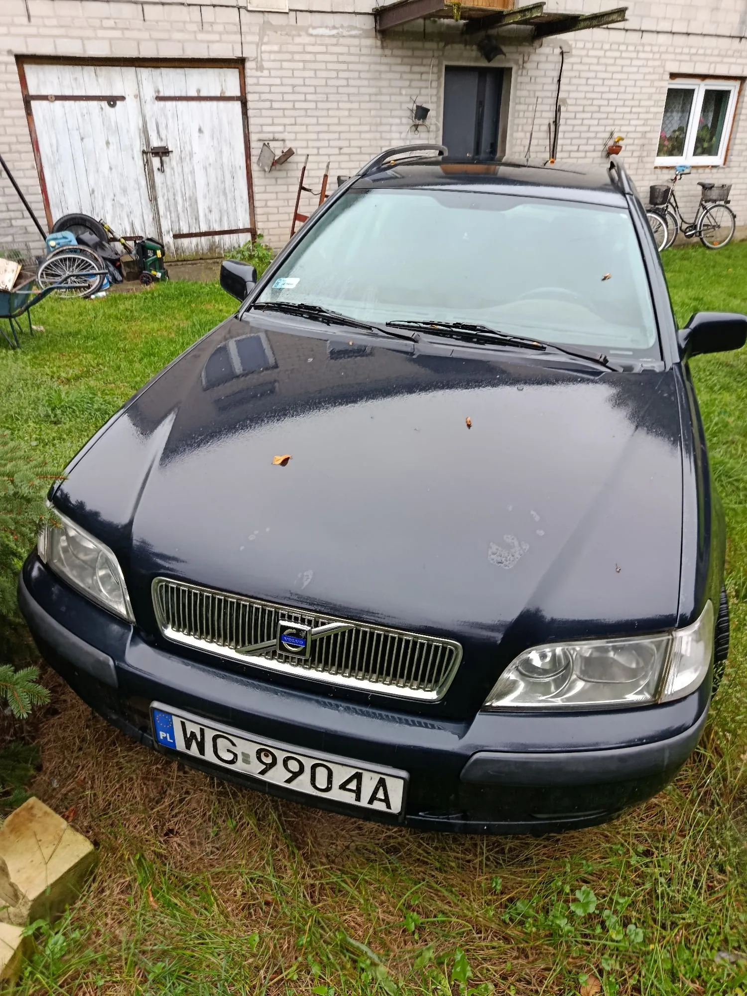 Czarny samochód marki Volvo zaparkowany na trawie przed białym budynkiem z cegły. Na przednim zderzaku znajduje się polska tablica rejestracyjna o numerze WG 9904A. W tle widoczne są białe drzwi garażowe, drzwi wejściowe oraz rower oparty o ścianę.