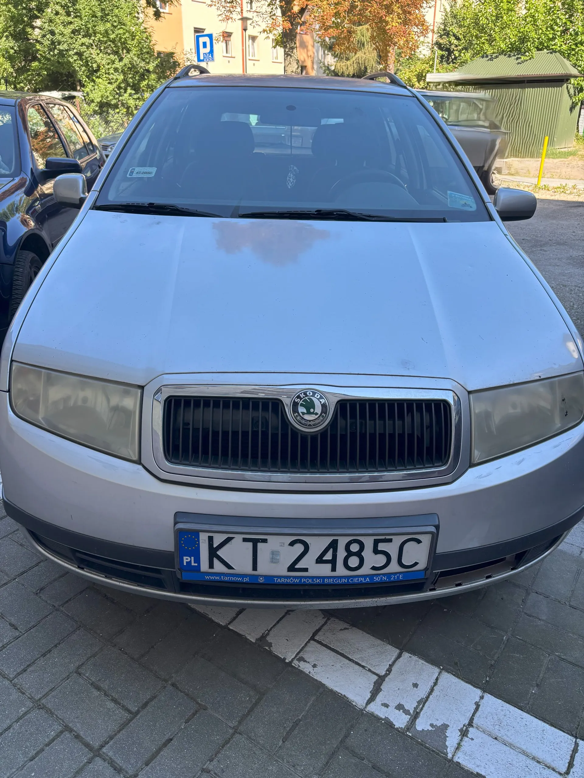 Srebrny samochód marki Škoda zaparkowany na kostce brukowej. Widoczny jest przód pojazdu z maską, reflektorami, grillem z logo marki oraz polską tablicą rejestracyjną o numerze KT 2485C. W tle widać inne zaparkowane samochody, drzewa oraz fragment budynku.