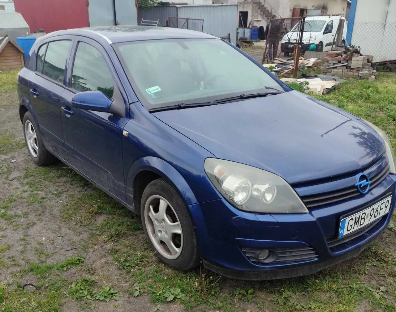 Niebieski samochód marki Opel z tablicą rejestracyjną GMB 96F9 (PL) zaparkowany na terenie z trawą i żwirem. W tle widoczne są budynki, biały furgon, materiały budowlane (cegły, deski), ogrodzenie i niewielka drewniana budowla. Na przedniej szybie samochodu znajduje się niebieski naklejony znaczek.
