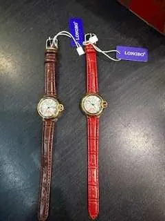 Dwa zegarki na ciemnej powierzchni. Górny z czerwonym paskiem, dolny z brązowym. Oba mają okrągłe, złote obudowy i białe tarcze. Do lewej strony zegarków przyczepione są niebieskie etykiety z białym tekstem: 'JORDAN' oraz 'JORDAN'S'.