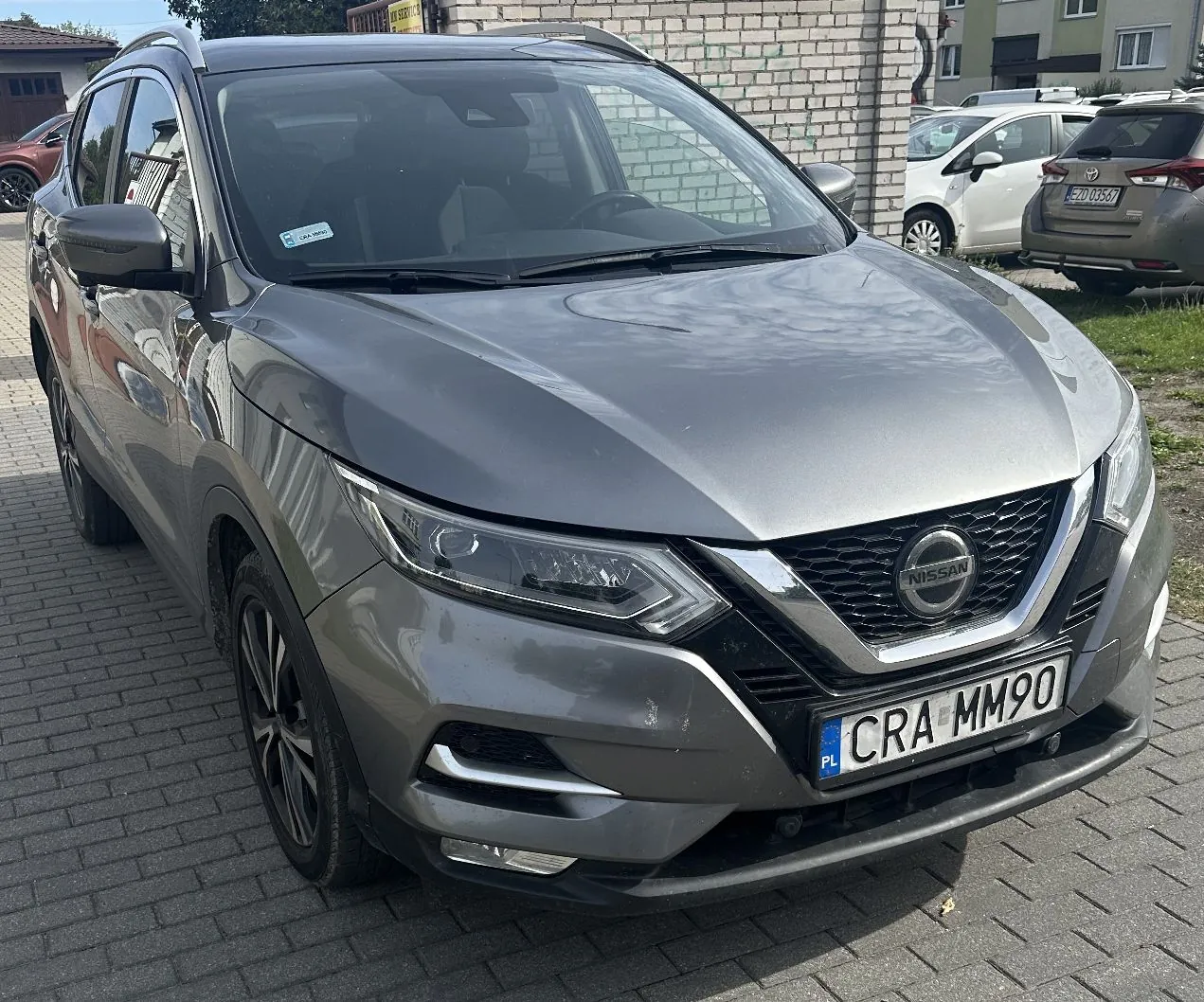 Srebrnoszary samochód marki Nissan Qashqai zaparkowany na kostce brukowej. Pojazd jest ustawiony przodem do widza, lekko pod kątem. Na przodzie widoczna jest czarna atrapa chłodnicy z logo Nissan oraz polska tablica rejestracyjna o numerze CRA MM90. W tle znajduje się biały mur z cegieł oraz dwa inne zaparkowane samochody: biały i szary.