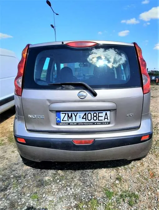 Tył srebrnego samochodu Nissan Note z widoczną tablicą rejestracyjną ZMY 408EA. Samochód stoi na żwirowym podłożu, w tle błękitne niebo z kilkoma chmurami oraz fragment białego pojazdu po lewej stronie. Na tablicy rejestracyjnej widoczny jest niebieski pas z napisem PL oraz dodatkowy napis pod tablicą: RENAULT FOLIA 20700 Myszków. W tle widoczne są latarnie uliczne.
