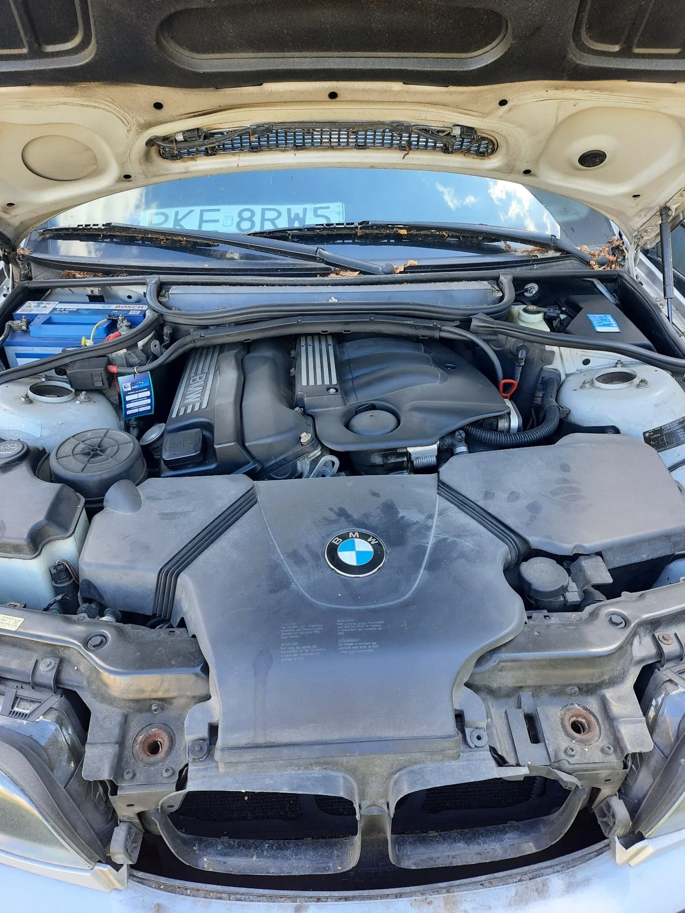 Widok z góry na otwartą maskę samochodu marki BMW, ukazującą komorę silnika. W centrum znajduje się czarna plastikowa osłona silnika z logo BMW. Po lewej stronie widoczny jest niebieski akumulator, a w tle, za przednią szybą, znajduje się tablica rejestracyjna o numerze PKF 8RW5.