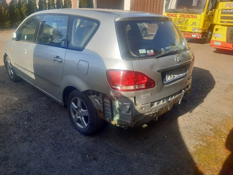 Zdjęcie przedstawia srebrny samochód osobowy marki Toyota z uszkodzonym tyłem, w tym zdeformowanym zderzakiem i częściowo uszkodzonym lewym tylnym światłem. Na tablicy rejestracyjnej widoczny jest numer 'PRE 455'. Na tylnej szybie znajdują się naklejki. W tle widoczne są dwa żółte pojazdy ratunkowe z napisami 'POMOC DROGOWA' oraz 'DAP', a także budynek z drewnianymi drzwiami i zielonymi krzewami po lewej stronie. Samochód stoi na podłożu z żwiru.