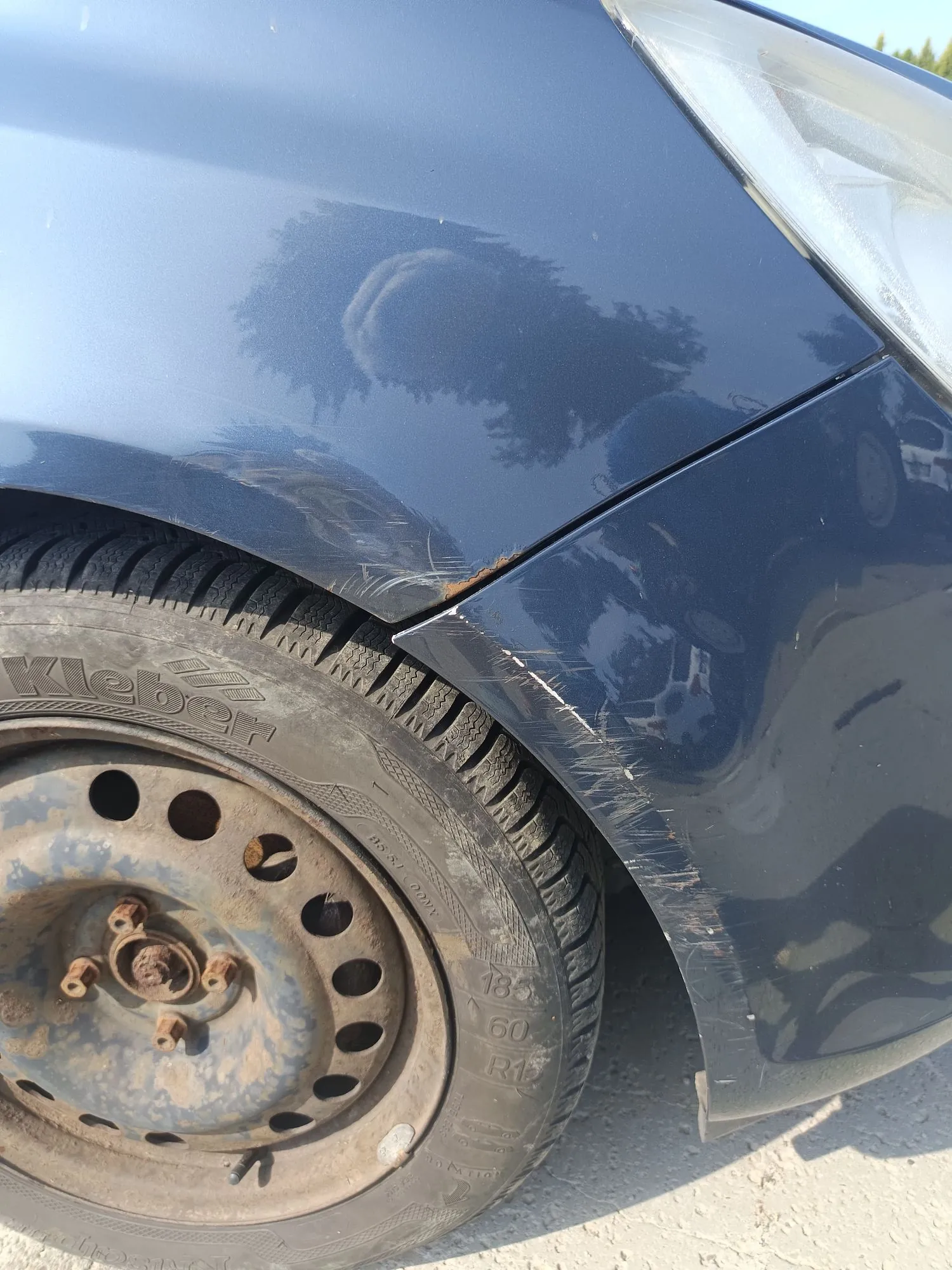 Zbliżenie na uszkodzony przedni prawy błotnik ciemnoniebieskiego samochodu. Metal jest mocno wgnieciony i pognieciony nad przednim kołem, z widocznymi zarysowaniami lakieru. Widoczna jest również część opony i stalowej felgi.