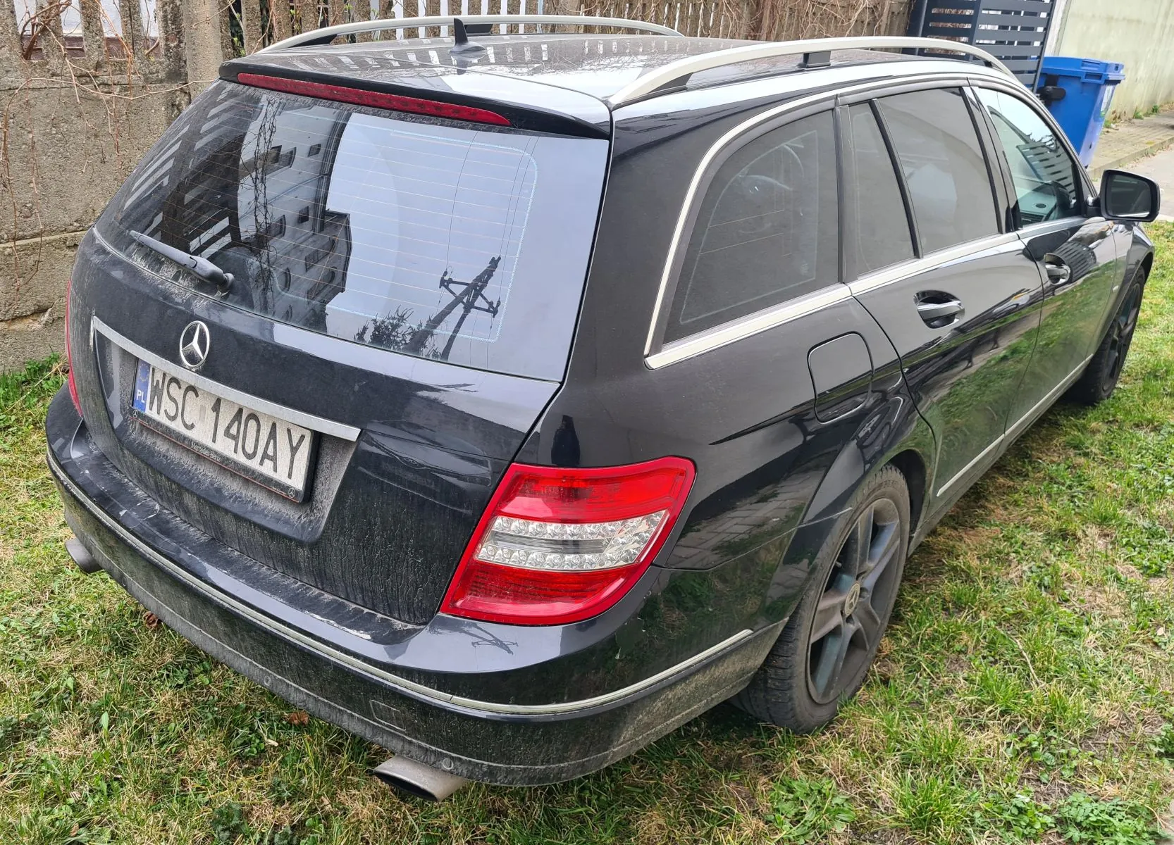 Czarny samochód osobowy typu kombi marki Mercedes-Benz zaparkowany na trawie. Widoczny tył pojazdu z tablicą rejestracyjną WSC 140AY oraz oznaczeniem PL. Samochód posiada dachowy bagażnik, przyciemniane szyby i czerwone światła tylne. W tle betonowa ściana z pnąciami, niebieski pojemnik na śmieci oraz metalowe ogrodzenie. Samochód jest lekko zabrudzony, szczególnie dolne części nadwozia.