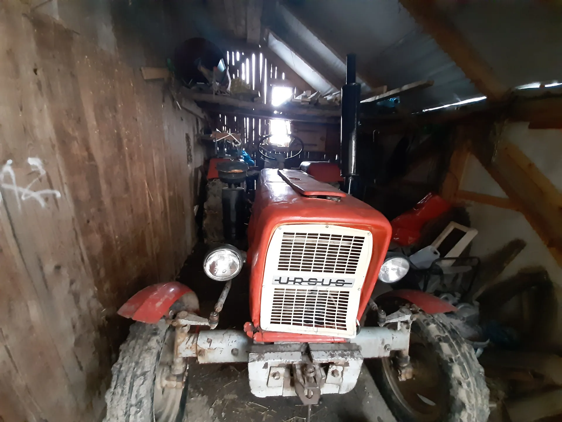 Czerwony traktor marki URSUS umieszczony w drewnianym garażu lub stodole. Traktor posiada białą kratę chłodnicy z widocznym napisem "URSUS", dwa reflektory oraz czerwone elementy nadkoli. W tle widoczne są drewniane ściany, dach z blachy, narzędzia i różne przedmioty przechowywane na półkach oraz na podłodze. Na lewej ścianie znajduje się biały napis lub znak. Oświetlenie pochodzi z otworów w dachu i ścianie.