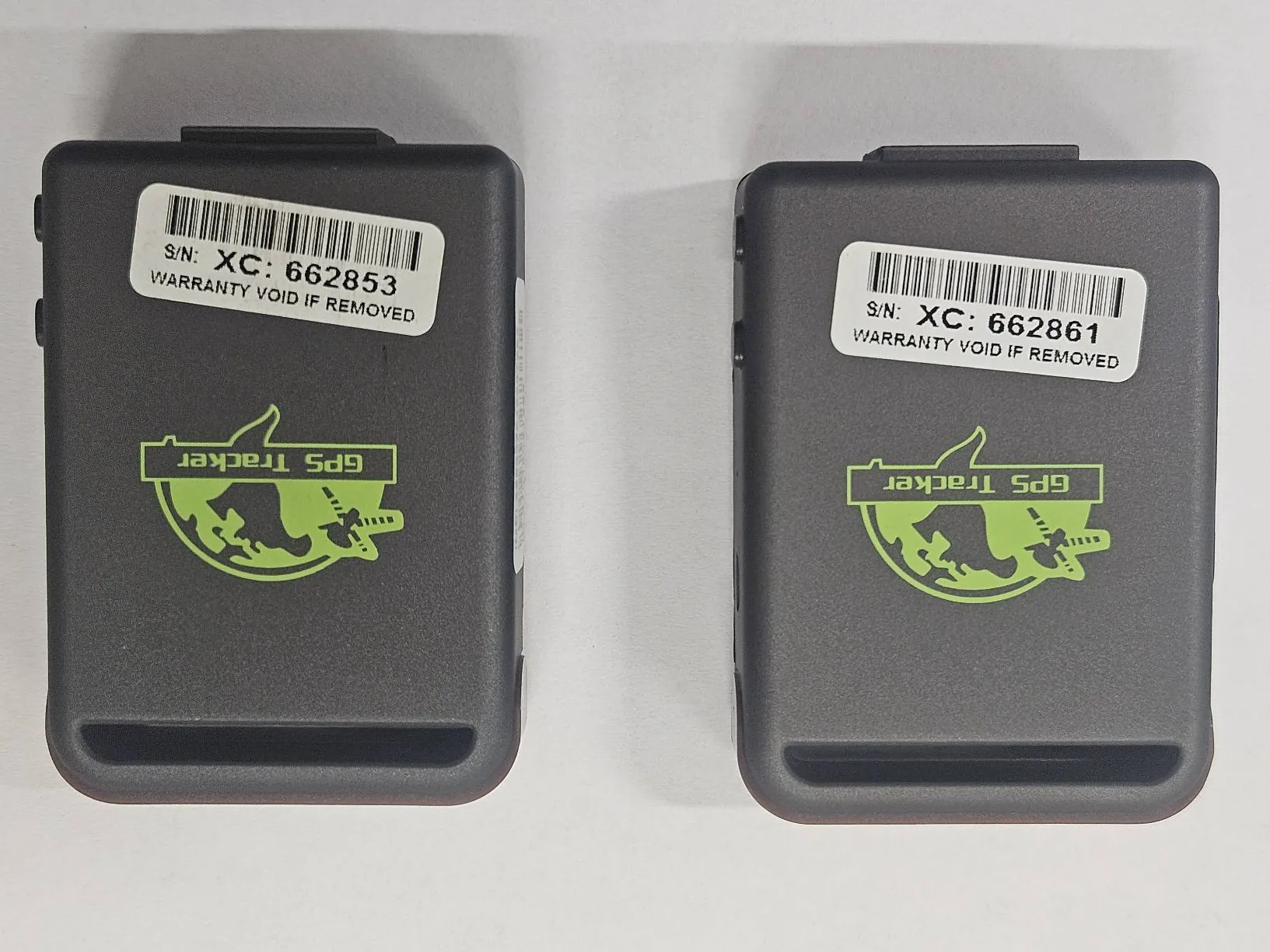 Dwa czarne urządzenia GPS Tracker ustawione obok siebie na białym tle. Każdy posiada zielony nadruk z globusem, satelitą i napisem 'GPS Tracker'. Na dolnej części każdego urządzenia znajduje się biała etykieta z kodem kreskowym oraz tekstem 'WARRANTY VOID IF REMOVED', 'S/N: XC: 662861' (po lewej) i 'S/N: XC: 662853' (po prawej).