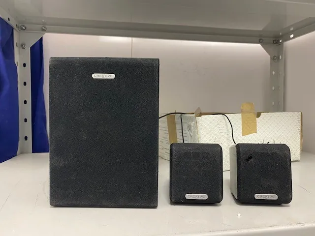 Trzy czarne głośniki marki CREATIVE ustawione na białej półce. Po lewej stronie znajduje się większy głośnik, a po prawej dwa mniejsze. Wszystkie mają widoczny napis 'CREATIVE' na przedniej części. W tle widoczne są metalowe elementy półki, niebieska tkanina po lewej stronie oraz kartonowe pudełko z taśmą klejącą. Półka ma metalowe wsporniki z otworami.
